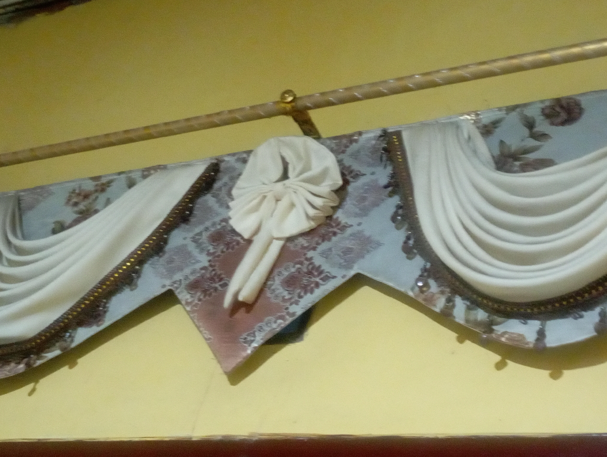 The image shows a valance, likely made of fabric, hanging from a golden rod attached to a light yellow wall. The valance is a decorative piece that hangs across the top of a window or doorway. It features a patterned fabric in shades of blue and maroon with floral motifs, and is embellished with white fabric folds and a cream-colored folded cloth centerpiece. White drapes hang from the sides of the valance. The valance is trimmed with a decorative braid of gold and dark beads. This scene is located in a domestic setting, specifically the city of Maos Kidul, Indonesia.