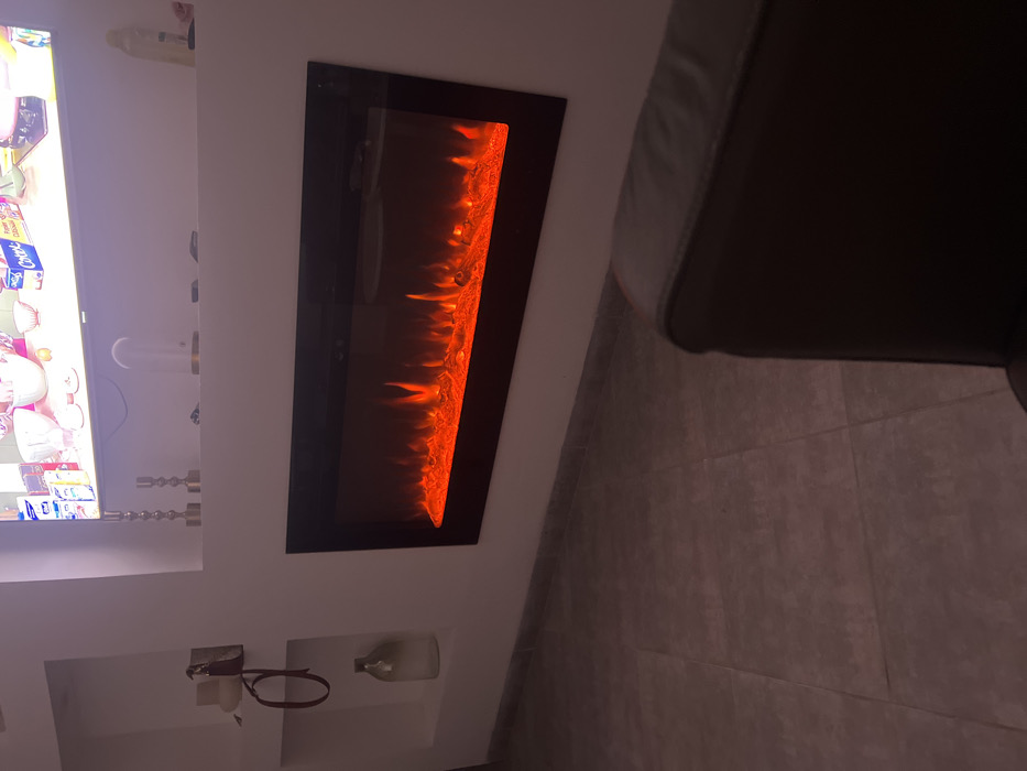 The image shows a living room with a modern electric fireplace set into a white wall. A television is mounted to the left of the fireplace. Decorative candles sit on a shelf below the TV, and a small shelf below the fireplace holds a glass bottle and a dog leash. The floor is tiled. A dark-colored piece of furniture, possibly a couch or chair, is on the right side of the frame.