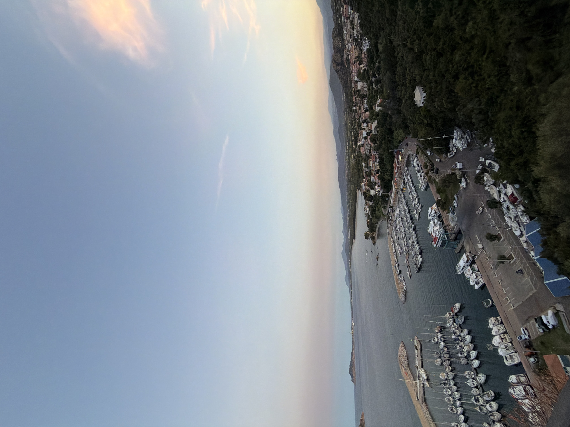 This is an aerial photograph of the Santa Maria Navarrese marina in Santa Maria Navarrese, Italy, taken during twilight. The sky is a gradient of light blue to pale pink, with a few wispy clouds illuminated by the setting sun.
The marina is filled with numerous boats of various sizes, predominantly white sailboats and motor yachts, docked in neat rows. Some larger vessels, including what appears to be a ferry or cruise ship, are also present. The water in the marina is calm and dark.
Adjacent to the marina is a town with buildings featuring red-tiled roofs, nestled among green trees. Hills and mountains form the backdrop of the town and the coastline. A road winds through the scene, connecting different areas and passing by parking lots with a few cars visible. The overall impression is of a serene coastal town at dusk.