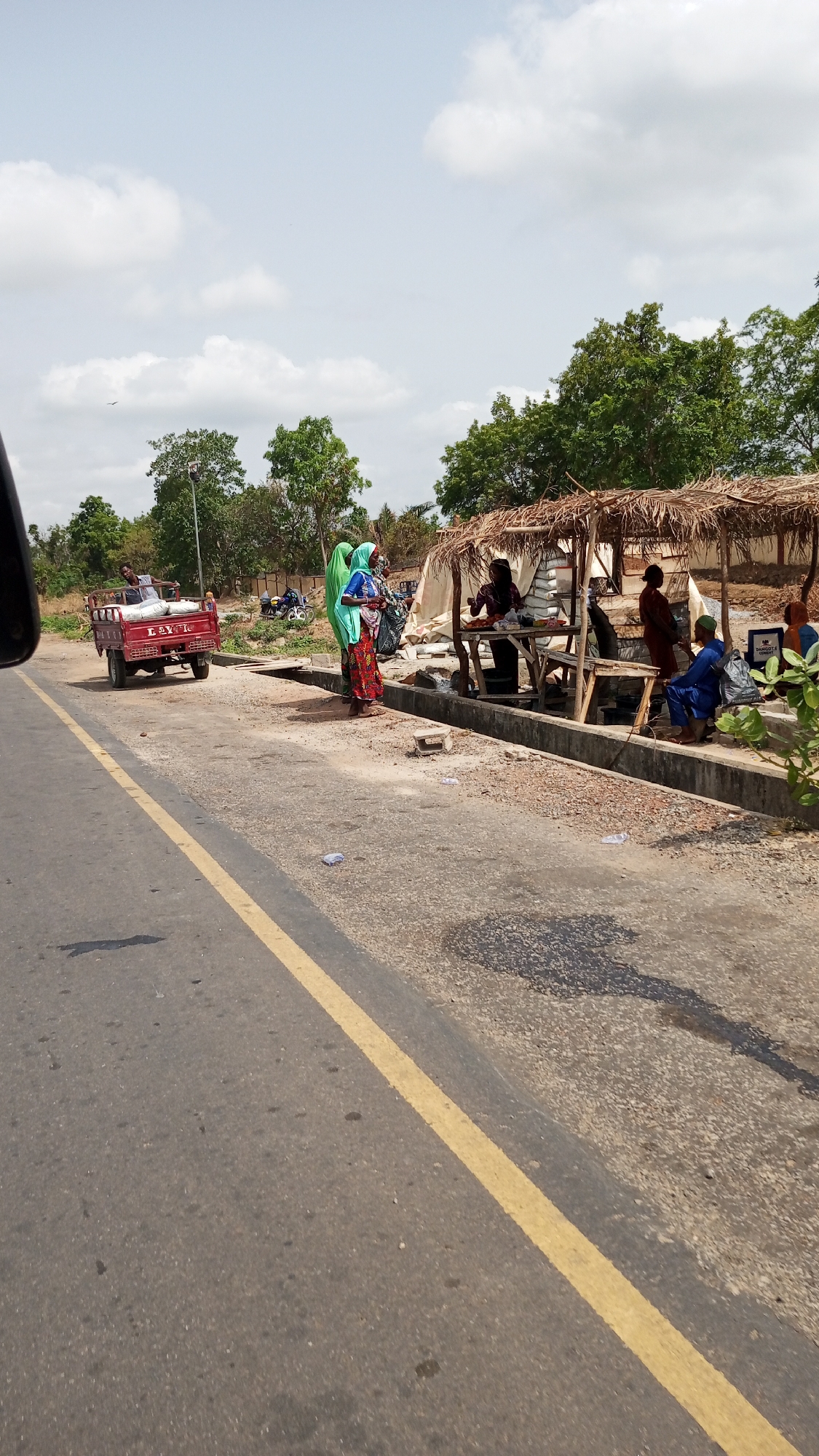 The scene shows a roadside setup on the outskirts of Zango-ndaji, Nigeria, next to a paved road with a yellow dividing line. On the right side, a makeshift shelter with a thatched roof provides shade for several individuals engaged in what appears to be commerce or social interaction. At least three people are under the shelter, with one standing and others seated. In front of the shelter, two women wearing traditional attire, including head coverings, stand near a raised concrete curb. To the left, a red motorized cart with cargo on the back is positioned off the road, with a person on the cart. The environment includes trees and vegetation in the background, suggesting a rural or semi-rural setting. The sky is overcast with clouds. There is debris scattered along the roadside, adding to the unkempt appearance of the area.