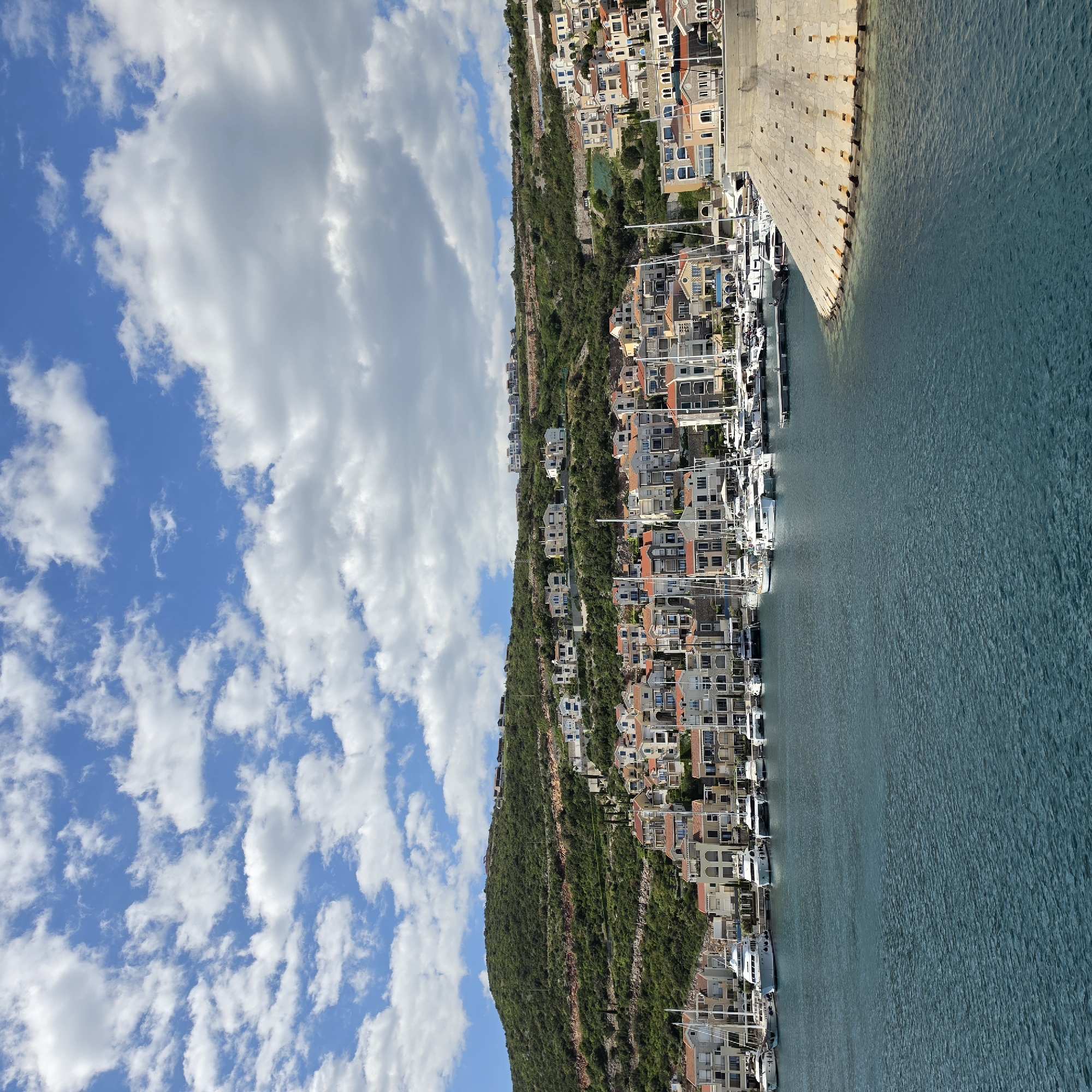 The image captures a picturesque coastal scene, likely in Radovići, Montenegro, as indicated. The perspective is from the water or a raised vantage point overlooking a bay.
The upper portion of the frame is dominated by a bright blue sky, adorned with scattered, fluffy white cumulus clouds, suggesting fair weather.
Below the sky, on the left side of the frame, a lush, verdant hillside rises, covered in dense green vegetation. Numerous residential buildings and houses are constructed along this sloping terrain, seemingly integrated into the landscape. These structures exhibit a variety of architectural styles, with some featuring light-colored facades and others adorned with traditional terracotta roofs, characteristic of Mediterranean coastal regions. Many of these homes appear to be multi-story and feature balconies or terraces offering views of the water.
Along the base of the hillside and extending into the middle ground, a bustling marina is visible. A multitude of boats and yachts, predominantly white, are neatly docked along the shore, their masts and rigging creating a dense pattern. The marina appears to be well-maintained, with walkways and docking facilities.
To the right side of the image, a significant body of calm, turquoise-blue water stretches out. The surface of the water shows gentle ripples, reflecting the clear sky. A large, robust concrete wall or breakwater forms a strong boundary between the open water and the inner harbor area where the boats are moored. This wall is textured and appears weathered, with dark square indentations or features along its length.
The overall impression is one of a vibrant yet serene coastal settlement, thriving between the verdant land and the sparkling sea, under a bright daytime sky. There are no visible people or text in the image.