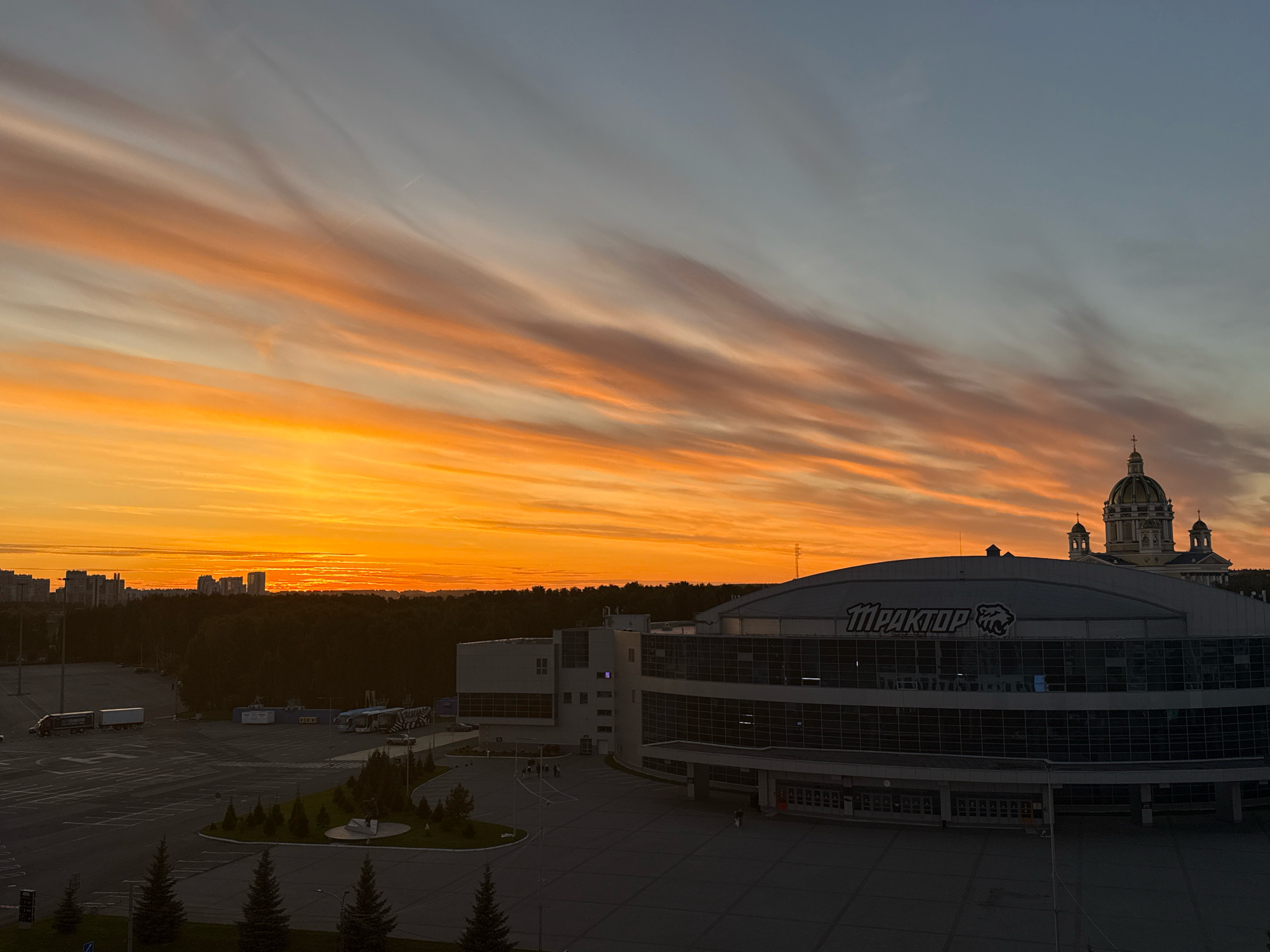 The image captures a sunset over the city of Chelyabinsk, Russia. The sky is filled with dramatic, wispy clouds painted in vibrant shades of orange, yellow, and pink, transitioning to a soft blue at the top. In the foreground, a large, modern sports arena with the prominent "ТРАКТОР" logo and a stylized panther is visible. The arena features a dark, curved facade with large glass windows reflecting the twilight sky. To the left of the arena, there is a large, paved parking lot with several vehicles, including trucks and buses. In the middle ground, a dense line of dark trees separates the parking lot from the city skyline. Several buildings, including apartment complexes, are visible in the distance under the colorful sky. To the right of the arena, a large, ornate building with a golden dome and several smaller domes, resembling a church or cathedral, stands prominently against the sky. The overall scene depicts an urban landscape at dusk, with a focus on the striking natural beauty of the sunset juxtaposed with man-made structures.