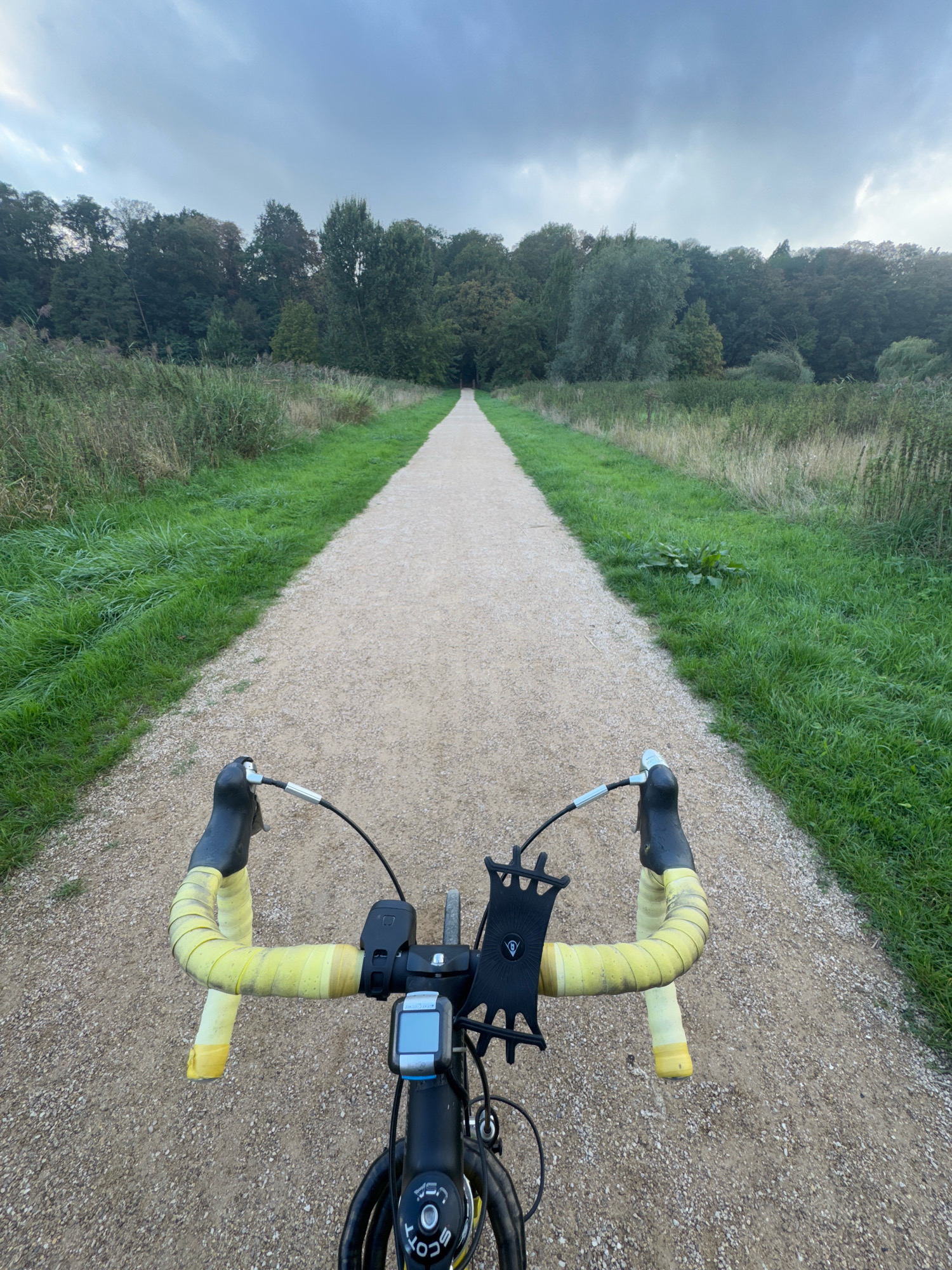The image is taken from the perspective of someone riding a bicycle on a gravel path. The path stretches into the distance, disappearing among trees on either side. The vegetation consists of tall grass and dense trees with green foliage. The sky is overcast with a cloudy, grey hue. On the handlebars of the bicycle, yellow handlebar tape is visible, along with a black phone mount and a bicycle computer. The bicycle itself appears to be a road or gravel bike with black components. The scene is set outdoors, in a natural or park-like environment. The location is not specified as Cleves, Germany, nor is there any indication of the city within the image.