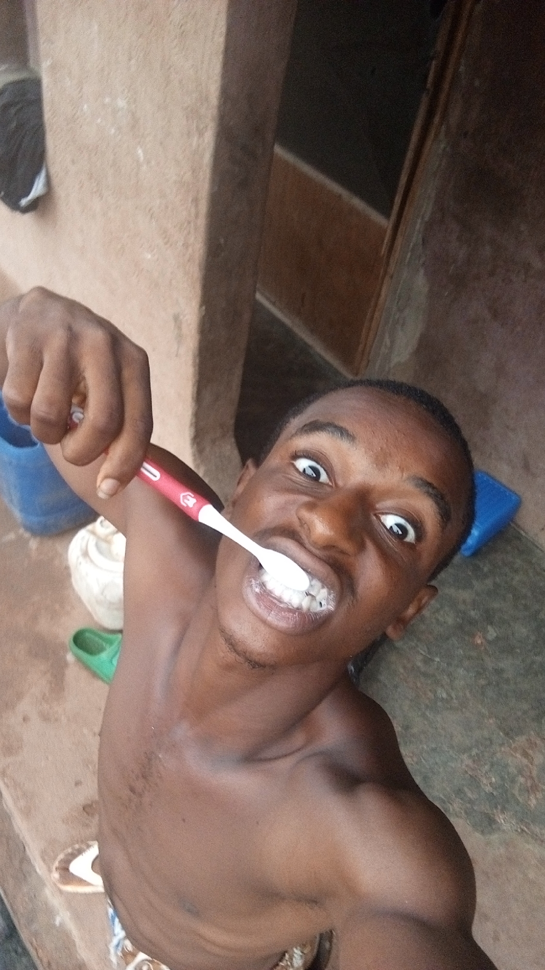 A shirtless young man is brushing his teeth with a red and white toothbrush, looking directly at the camera with wide eyes. He is standing outside, likely in a residential area in Onitsha, Nigeria. In the background are walls made of mud, and a doorway. There is a blue bucket, green sandal, and white and brown flip-flop visible on the ground.