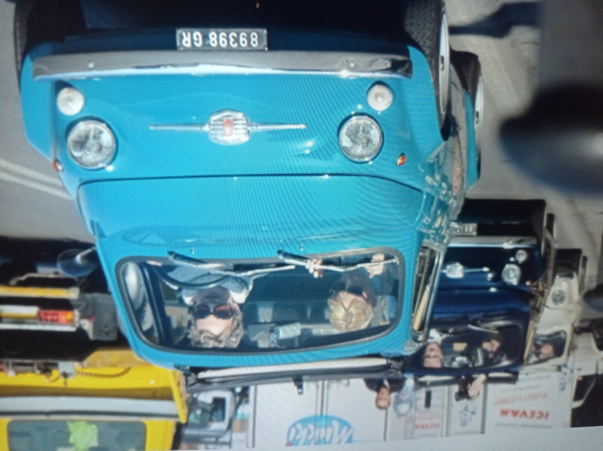 The image depicts a street scene in Mukachevo, Ukraine. The foreground features a bright blue Fiat 500 with two women inside. The driver, wearing sunglasses, is visible behind the steering wheel. The passenger, also wearing sunglasses, is to her left. The car's license plate reads "89398 GR". Behind the blue Fiat, a dark-colored car is partially visible. Further in the background, a white car and a dark blue car are parked. A yellow truck is also present. The environment appears to be a sunny day in a city.