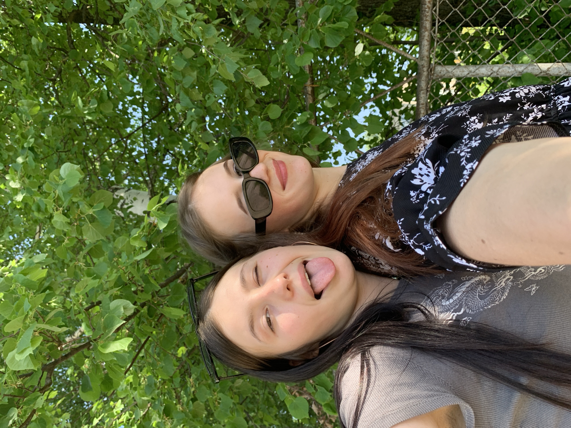 Two young women are posing for a selfie. The woman on top is wearing sunglasses and a black and white floral top. The woman on the bottom is sticking her tongue out and is wearing glasses and a gray shirt with a dragon design. They are positioned under a tree, with leaves and branches visible in the background. A metal fence is also visible in the background. The location is most likely in a park, and is in the city of Фоки, Russia.