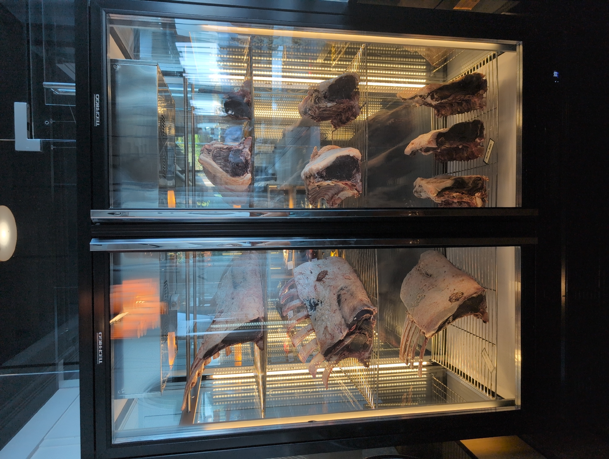 This is a full-frame, eye-level, medium shot of a double-doored, glass-fronted meat aging refrigerator. The refrigerator is predominantly black with a silver trim on the doors. Inside, on multiple metal shelves, are several large cuts of raw, dry-aged beef. The beef exhibits significant marbling and is predominantly a dark red, raw color with a greyish-white outer layer of fat and connective tissue.
The left side of the refrigerator contains three large cuts of beef. The top cut is a thick loin, the middle is a rack of ribs, and the bottom is another substantial piece of beef, possibly a sirloin or T-bone. The right side of the refrigerator has three shelves. The top shelf has two cuts of beef, the middle shelf has two more substantial cuts, and the bottom shelf has three smaller cuts. The interior of the refrigerator is illuminated by vertical rows of warm-toned LED lights behind the shelves.
The refrigerator is situated in an indoor environment with a dark ceiling. Reflections on the glass doors suggest an outdoor scene with windows and daylight. A digital display showing "39" is visible on the lower right corner of the refrigerator door. A label is present on the bottom shelf of the left side, and another label is visible on the bottom shelf of the right side. The text on these labels is illegible in the given resolution. The overall scene suggests a restaurant or butcher shop specializing in dry-aged beef.
The location is identified as Opatija, Croatia. Specifically, the image appears to have been taken at the restaurant Roko, located at Ul. Maršala Tita 106, 51410, Opatija, Croatia.