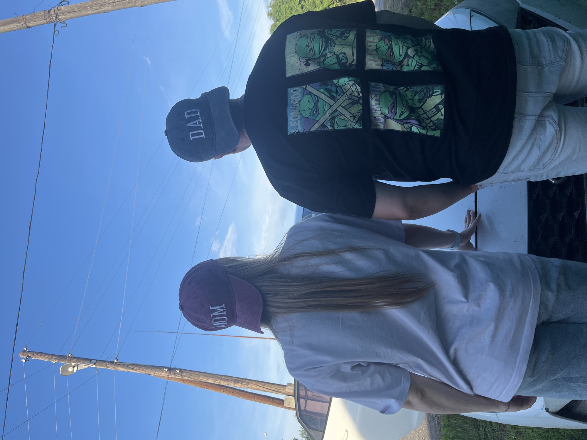 In Mal'tsevo, Russia, a couple stands facing away from the camera, leaning against the hood of a white car under a clear blue sky. The woman on the left wears a maroon baseball cap with "MOM" embroidered in white, and a light-colored t-shirt. The man on the right wears a dark gray baseball cap with "DAD" embroidered in white, and a black t-shirt featuring four panels of the Teenage Mutant Ninja Turtles. They are positioned in an outdoor setting with power poles and overhead wires visible in the background.