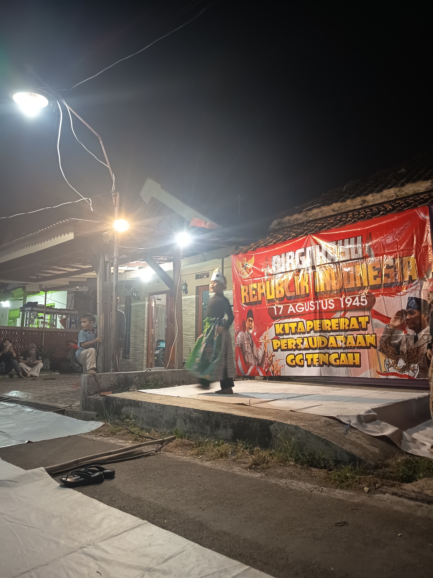 The image depicts an outdoor nighttime scene, possibly a community event. A large red banner with yellow lettering that reads "DIRGAHAYU REPUBLIK INDONESIA 17 AGUSTUS 1945 KITAPERERAT PERSAUDARAAN GG TENGAH" is prominently displayed. The banner features illustrations of historical figures and the Indonesian coat of arms. In front of the banner, a person in traditional attire, including a green skirt and a headpiece, is walking across a raised platform. Several people are seated and standing in the background, some appearing to be spectators. Overhead, a streetlamp illuminates the area, casting a bright light. The environment appears to be a street or a small gathering space, with a building structure visible behind the banner. The ground is paved, and there are some white tarps or coverings in the foreground. A pair of black flip-flops are also visible on the ground.
