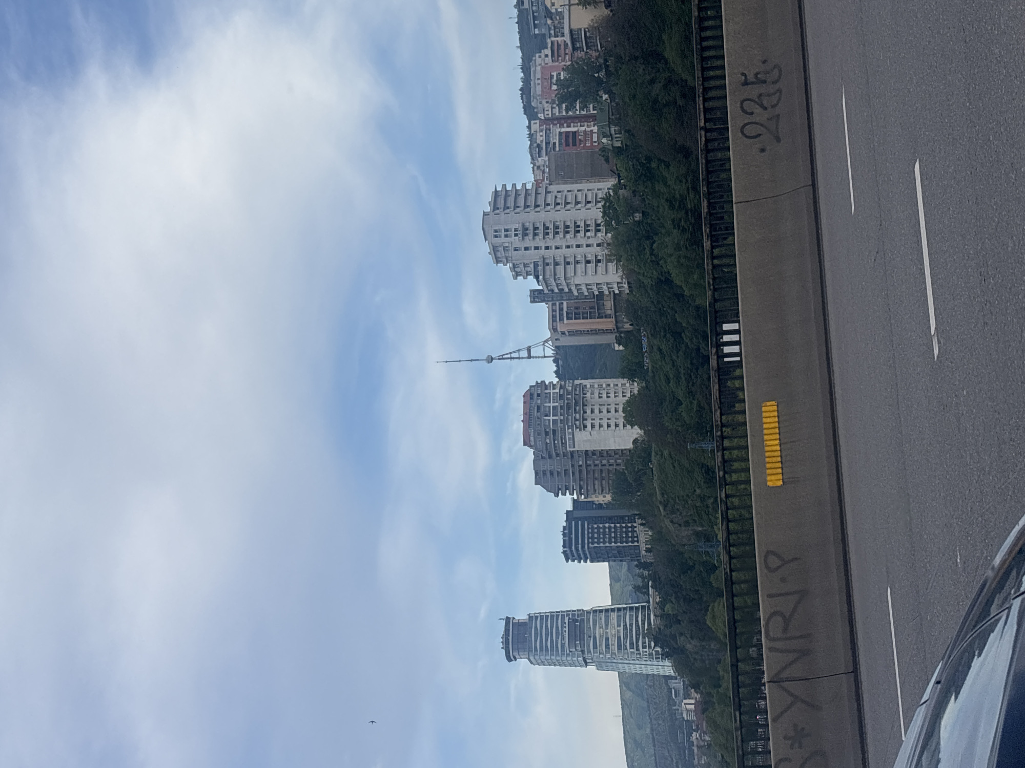 The image shows a view of the city of Tbilisi, Georgia, from a bridge or elevated roadway. The photograph is taken at an angle, with the city skyline visible against a cloudy blue sky. A tall television tower is visible among the buildings. The edge of the road is in the foreground, with graffiti and a yellow reflective panel. A portion of a car is visible at the bottom right of the image. The architectural style of the buildings suggests the city's urban environment.