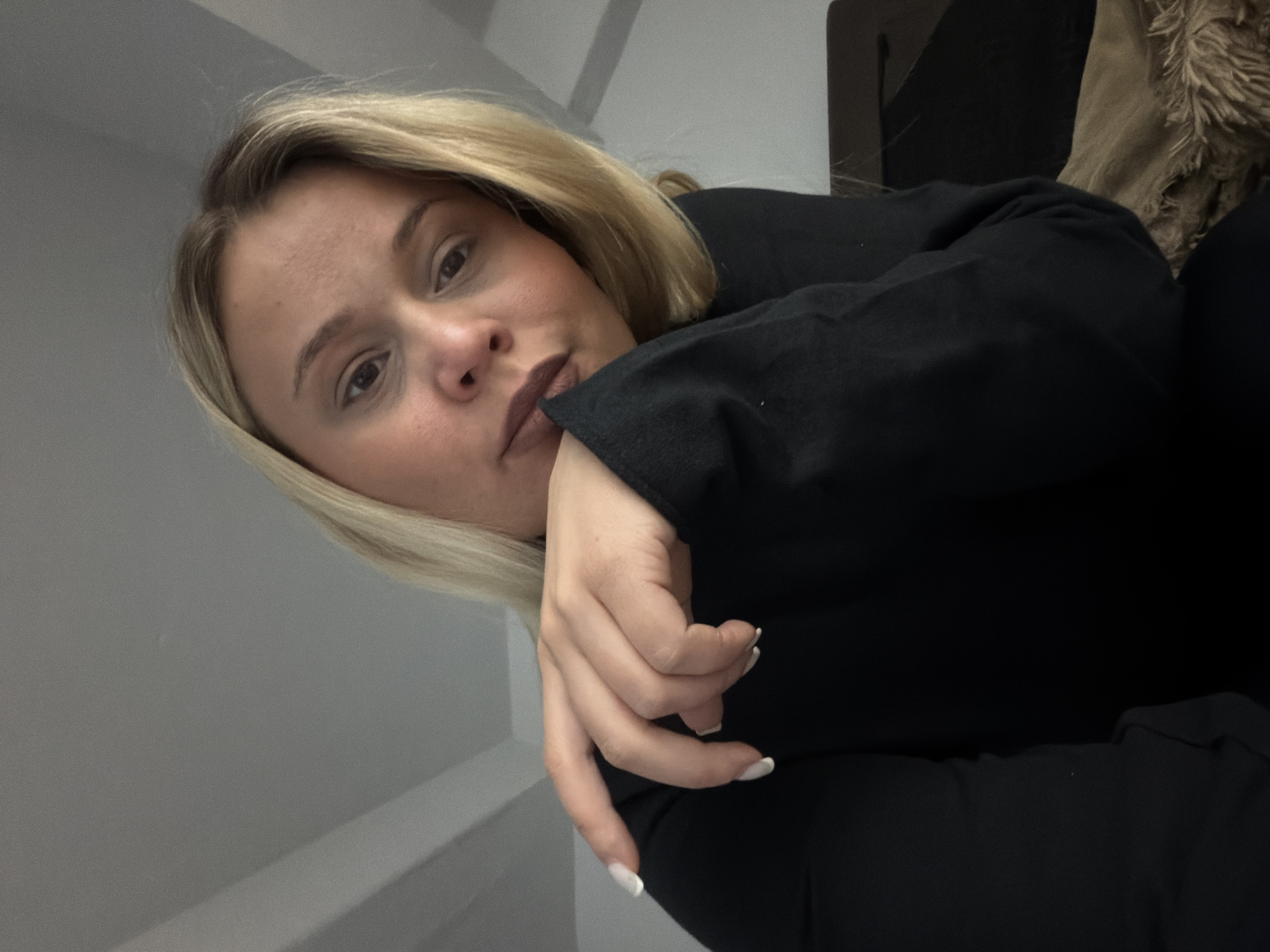 A woman with blonde hair and light skin is positioned in the foreground, looking toward the camera with a neutral expression. Her hand is near her face, with fingernails painted white. She is wearing a black garment. The background is a plain gray wall. The image does not contain information about the city of Sarapul, Russia.