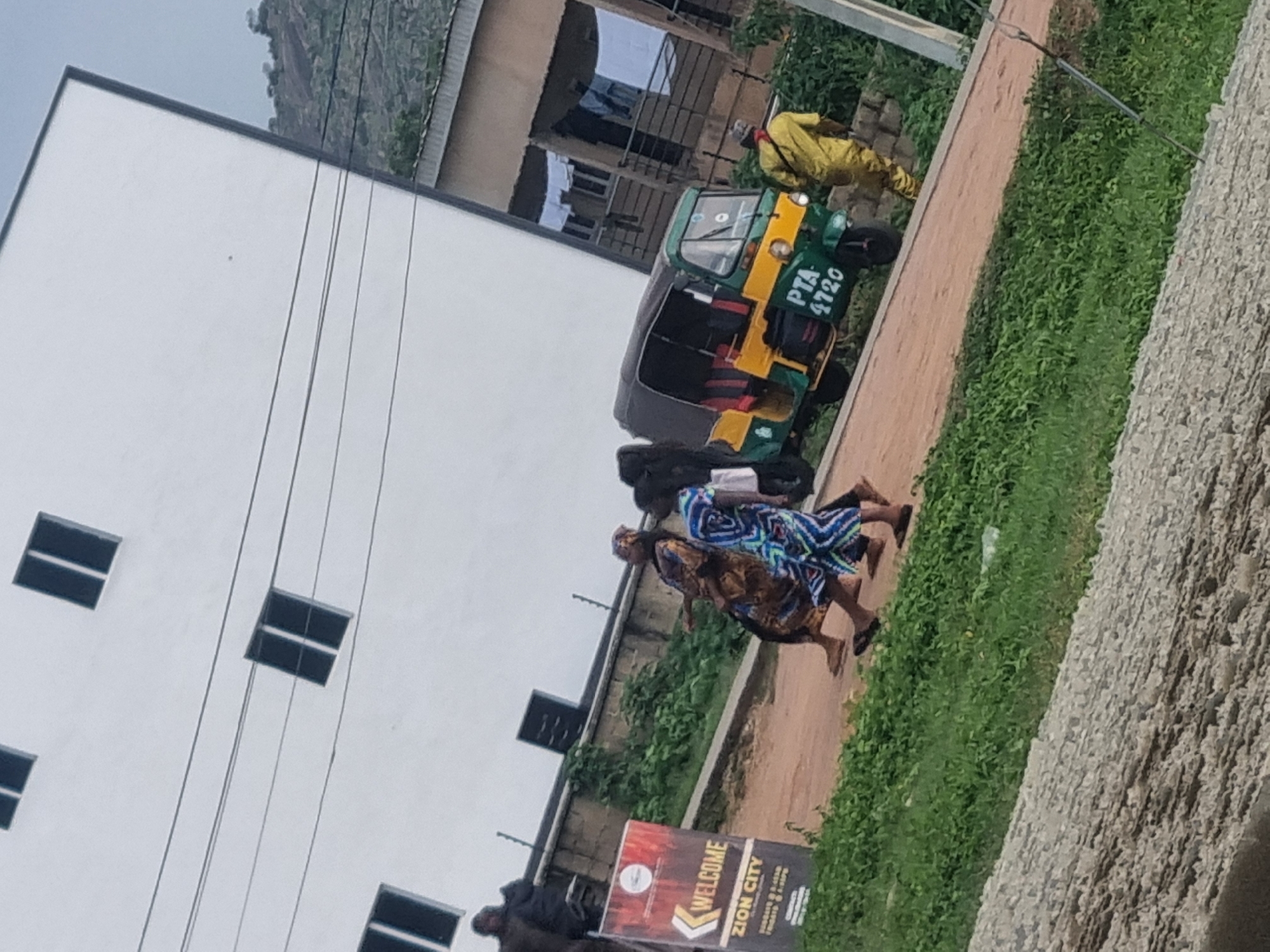 In Jos, Nigeria, a street scene unfolds with a modern white building dominating the background, its façade punctuated by dark, rectangular windows. Several utility wires crisscross the sky in front of the building. In the mid-ground, a group of people are walking on a dirt path. Two women, one in a brown and gold patterned dress and the other in a blue and white geometric print dress, are walking side-by-side. Behind them, another person in dark clothing is visible. To their right, a green and yellow auto-rickshaw, also known as a "Keke Napep," is parked. The auto-rickshaw has the license plate "PTA 4720" displayed. Further to the right, a man wearing a bright yellow uniform and a red bandana around his neck is walking away from the viewer. A "WELCOME ZION CITY" sign stands to the left of the path, with arrows pointing left and text detailing service times. The foreground is filled with lush green grass and rough, grey gravel. The overall atmosphere suggests an overcast day in a bustling urban environment.