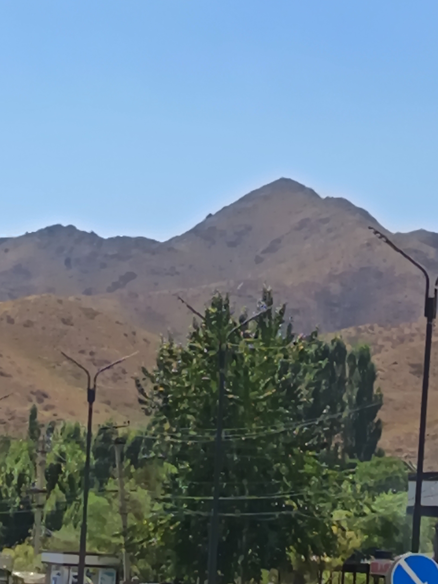 The image depicts a landscape scene with rolling, barren hills in the background under a clear blue sky. A prominent, cone-shaped mountain peak is visible in the center of the background. In the middle ground, a large, dense green tree with lush foliage dominates the scene. Two lampposts are visible in the foreground, one on the left and one on the right, with one on the left partially obscured by the tree. There are also power lines crisscrossing the middle ground. In the lower right corner, a blue and white circular traffic sign is partially visible. The overall impression is a rural or semi-rural environment with natural topography and some signs of infrastructure. No people or animals are discernible in the image.