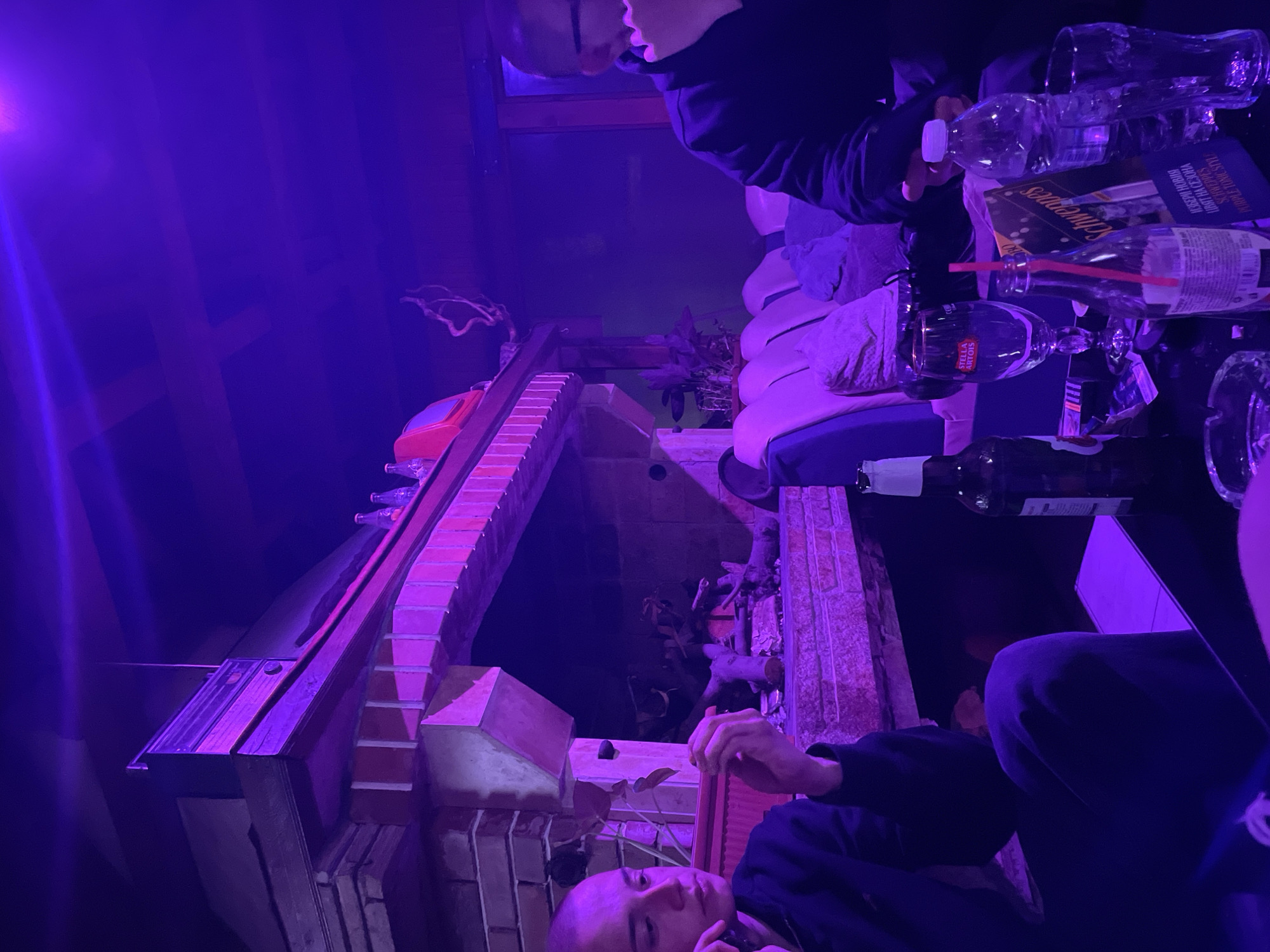 The image shows an indoor scene in Zlatograd, Bulgaria, illuminated by purple light. A brick fireplace and mantelpiece are visible. Two people are present. One individual is partially visible, holding a water bottle. The other individual, who is bald, is resting their head on their hand. A table cluttered with empty glasses, a bottle of Stella Artois, a Schweppes advertisement, and other objects is also visible. The environment appears to be casual, possibly a social gathering or a party.