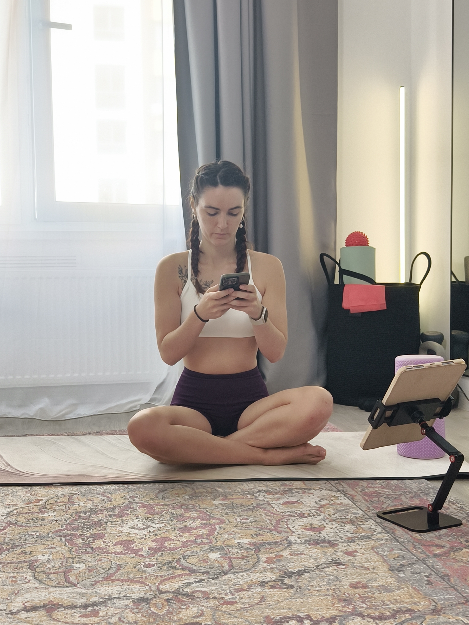 A woman with dark braided hair, wearing a white sports bra and dark purple shorts, is sitting cross-legged on a yoga mat in a room that appears to be in an apartment in Tyumen, Russia. She is looking down at her smartphone, which she holds in her hands. To her right, there is a tablet on a stand, a black storage bag containing yoga equipment, and a patterned rug covers the floor in front of the mat. A window with white curtains is visible on the left side of the image.