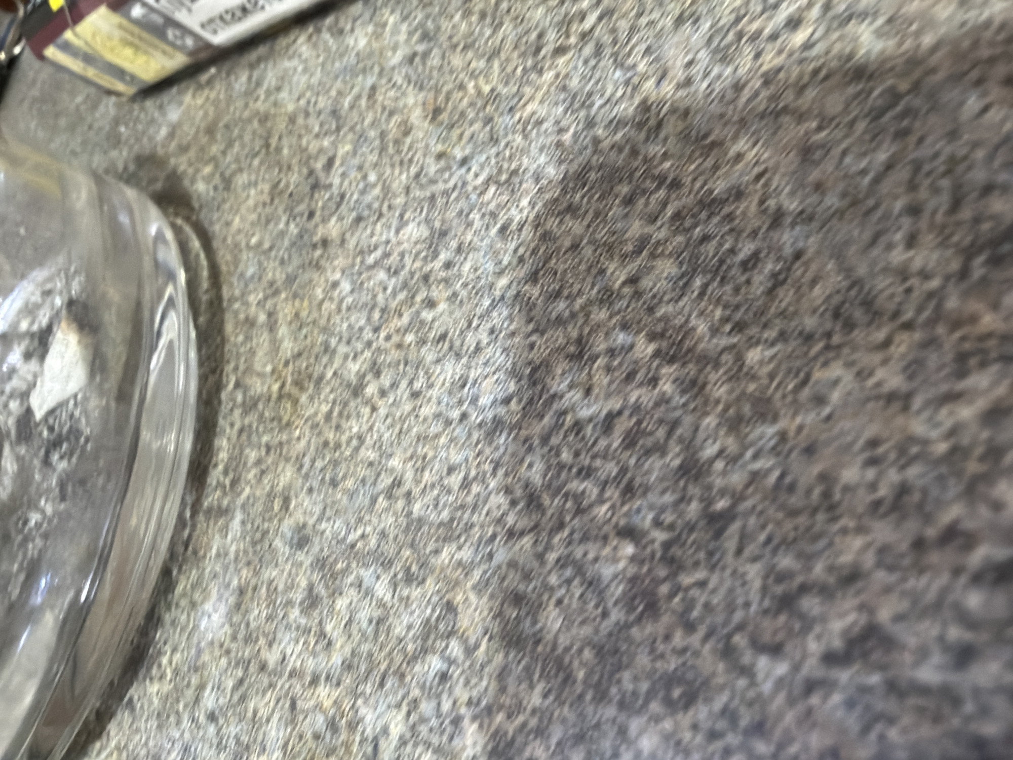 The image shows a close-up view of a countertop with a mottled grey and brown pattern. In the lower left corner, part of a clear glass object, possibly a dish or lid, is visible. A corner of a product box with yellow and white text is at the top left. The environment appears to be indoors. There is no information in the image to determine the location in Fenland District, United Kingdom. There are no people, actions, or interactions visible.
