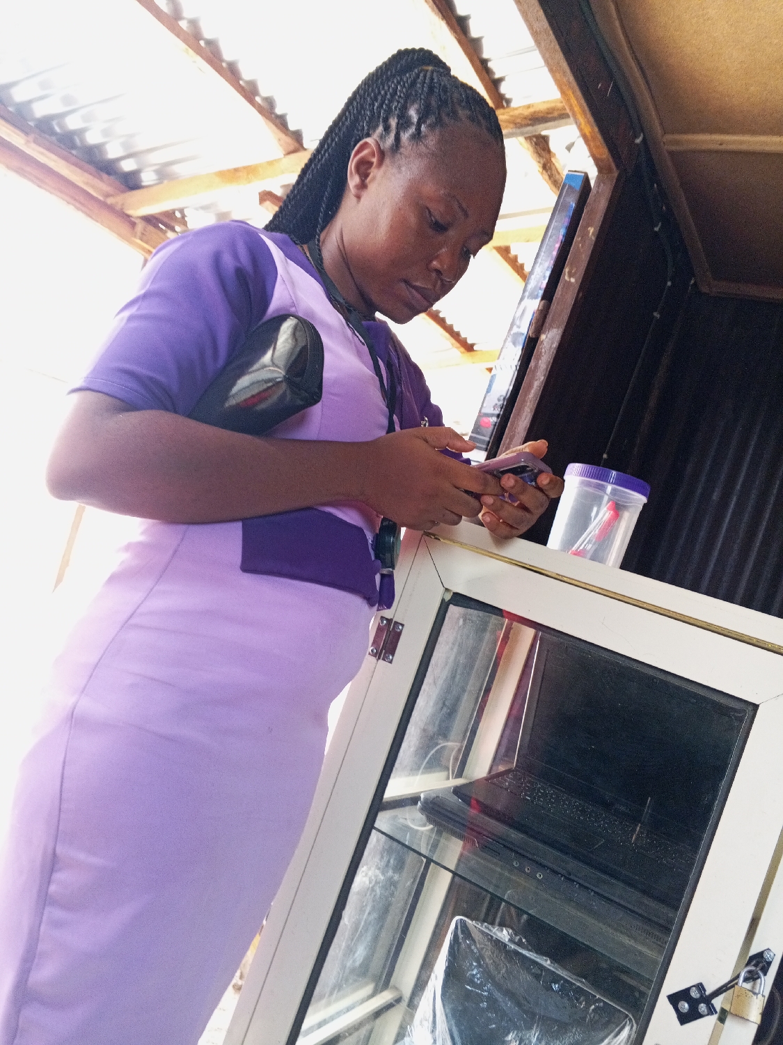 A young woman, likely in her 20s or 30s, is depicted indoors or in a semi-enclosed structure during the daytime in Adankolo, Nigeria. She has dark skin and her hair is styled in neat dark braids pulled back into a ponytail. She is wearing a light purple uniform or dress with darker purple accents on the short sleeves and neckline, and a black handbag is visible across her chest.
The woman is focused on her smartphone, holding it with both hands and looking down at its screen. She appears to be engrossed in an activity on the device.
To her right, a white-framed display cabinet with glass panels is visible. Inside the cabinet, a black laptop is placed on a shelf, and another electronic item wrapped in clear plastic rests on a lower shelf. A padlock is attached to the exterior of the cabinet, suggesting it is used for secure storage or display of merchandise. Next to her right hand, on the surface of the cabinet, sits a clear plastic container with a purple lid.
Above her head, a corrugated metal roof supported by wooden beams is visible, indicating a utilitarian structure, possibly a market stall or a small shop. The lighting is bright, with some areas appearing slightly overexposed, characteristic of daylight. There is no visible text in the image.