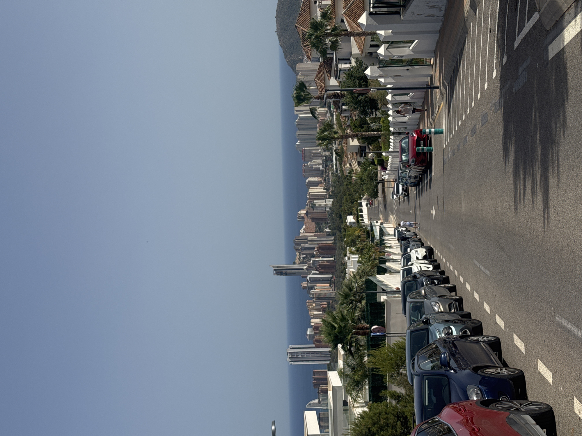 The image captures a street scene in Finestrat, Spain, overlooking a coastal cityscape. The foreground features a street lined with parked cars, predominantly on the right side of the road. A red car is prominently positioned on the left, while a row of other vehicles, including a dark blue hatchback, a silver sedan, and several white cars, are parked along the right curb, which is demarcated by white dashed lines. A man in a white t-shirt and shorts is walking away from the camera down the sidewalk on the right. The street itself is asphalt, with white lane markings visible.
In the midground, the view opens up to reveal a dense urban landscape characterized by numerous buildings and high-rise structures, likely residential and commercial properties, situated against the backdrop of the Mediterranean Sea. The architecture varies, with modern skyscrapers standing alongside older, lower-rise buildings with terracotta-tiled roofs. Palm trees and other greenery are scattered throughout the urban area, indicating a Mediterranean climate.
The background is dominated by the clear, pale blue sky and the vast expanse of the sea. To the right of the cityscape, a rolling, green-covered hill rises, with some structures visible on its slopes and summit. The overall impression is one of a sunny day in a coastal city with significant vertical development.