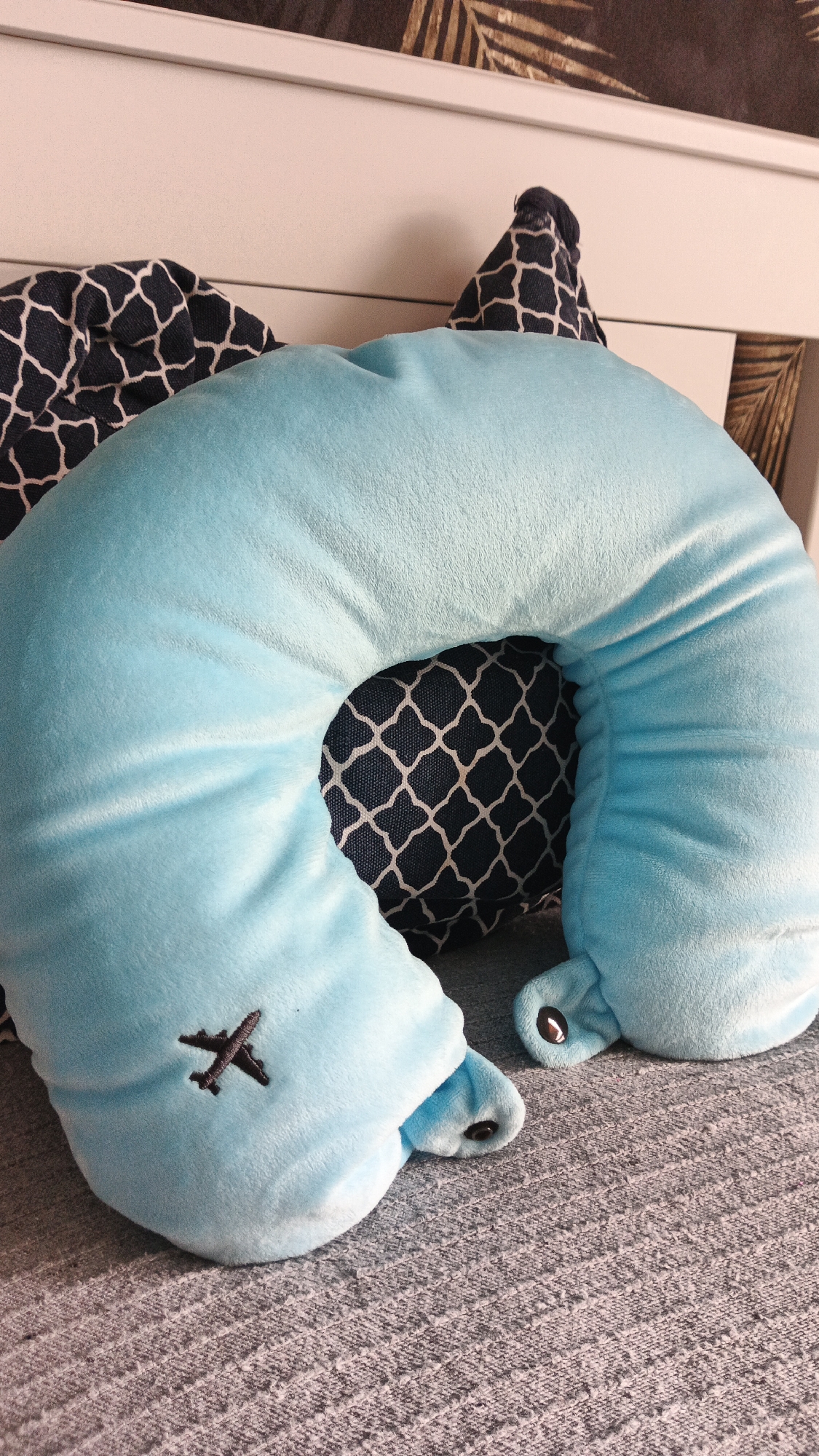 A light blue u-shaped pillow with a dark navy and white patterned pillow underneath rests on a grey textured surface. A small black airplane is embroidered on the left side of the u-shaped pillow. The scene appears to be indoors, possibly in a bedroom, given the presence of a headboard and decorative wallpaper in the background. The image does not provide any information about the city of Zielona Góra, Poland.