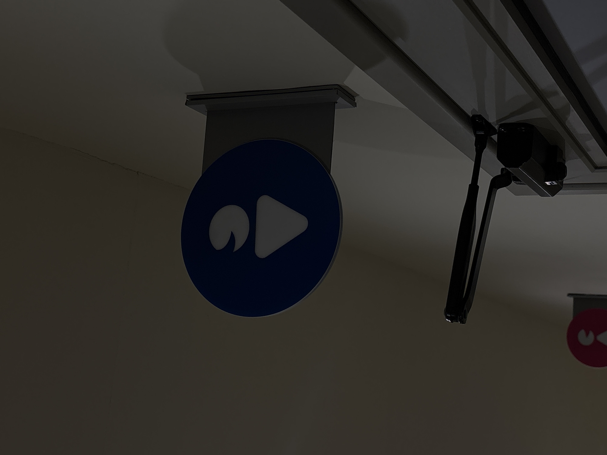 A low-angle, indoor shot depicts a blue, round sign with a white graphic consisting of a partial circle and a right-pointing triangle. The sign is suspended from the ceiling by a gray metal bracket. To the right of the sign, attached to the ceiling, is a black articulating arm and camera. In the background, another sign, red with white graphics, is visible. The location is suggested as Yerevan, Armenia. The room is dim, with light primarily reflecting off the ceiling.