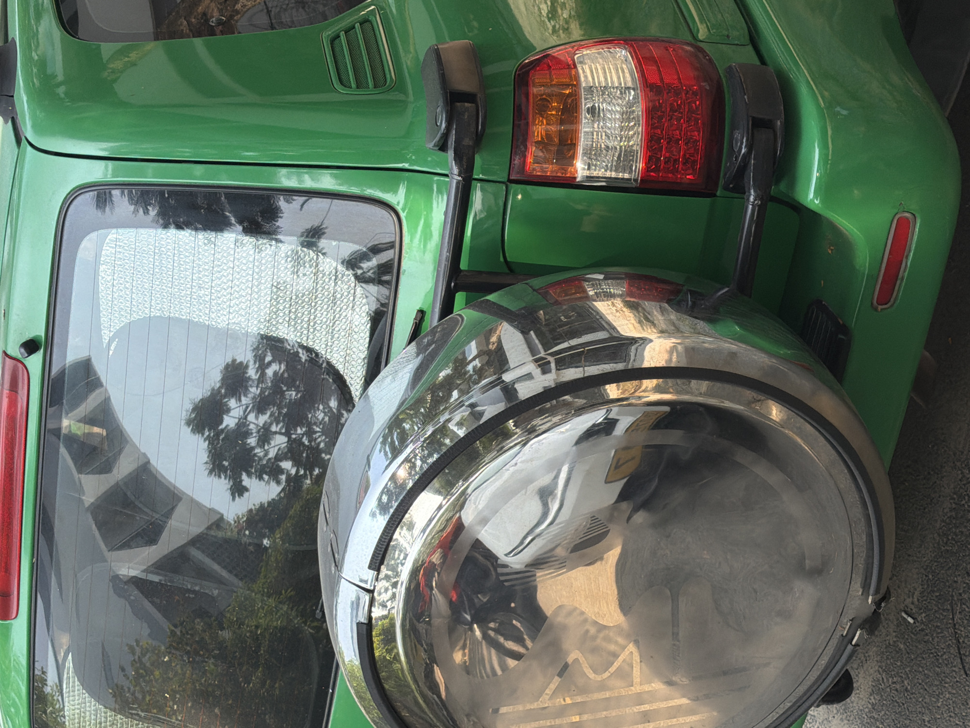 The image shows the rear of a green vehicle, potentially a car or SUV, in what appears to be Đà Nẵng, Vietnam. This is suggested by the reflection in the vehicle's rear window, which depicts a building structure characteristic of urban Southeast Asia, along with the sparse greenery and climate seen within the reflection. The vehicle has a spare tire mounted on the back, enclosed within a chrome tire cover. A taillight is visible, along with the black mounting brackets for the spare tire. The environment seems to be a city setting, likely either a street or parking area.