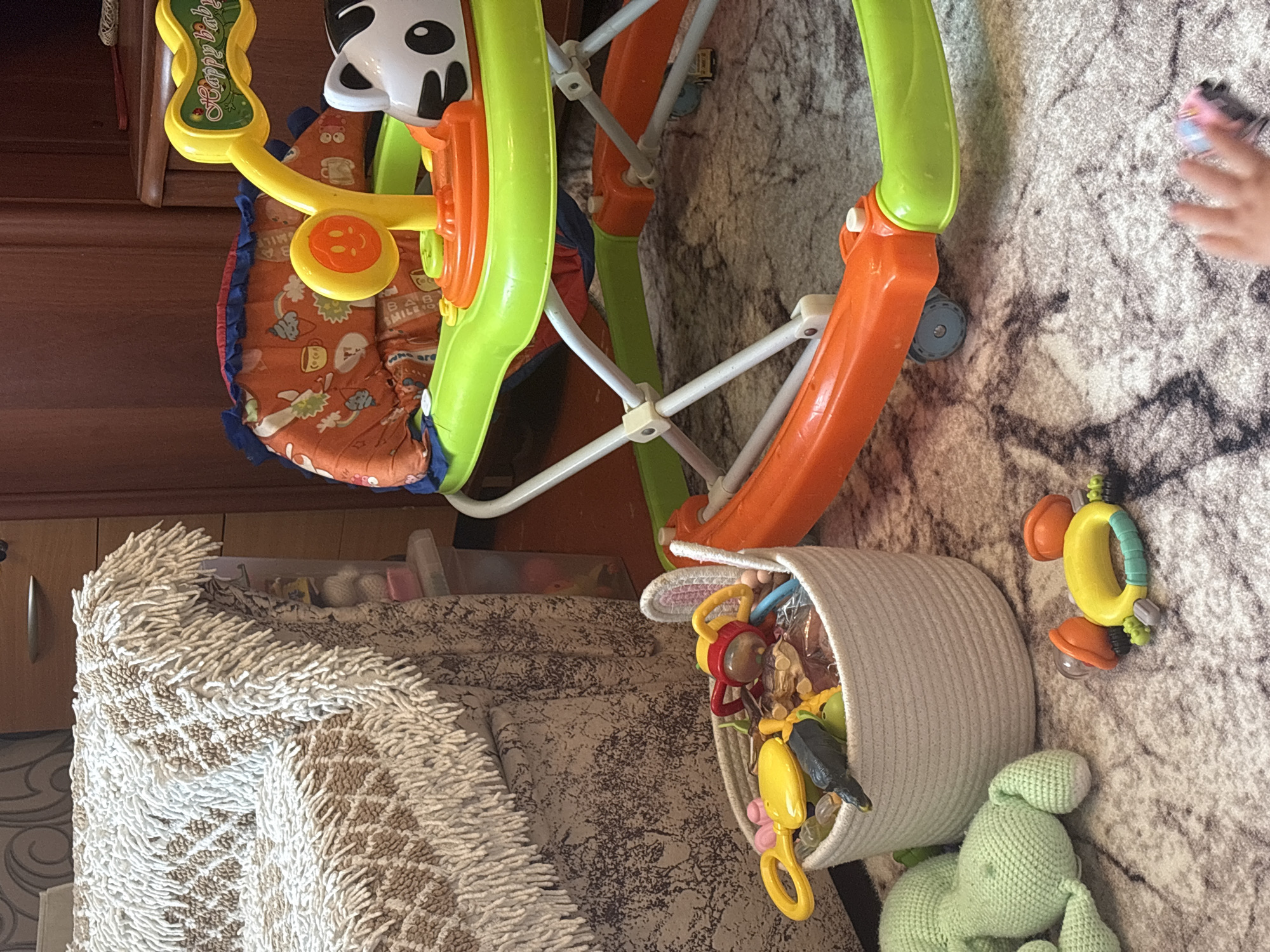 The image depicts a dimly lit room, likely a living area, with a focus on children's toys and furniture. In the foreground, a white woven basket overflows with various baby toys, including rattles, teethers, and a small dark toy animal. To the left, a large, light green crocheted stuffed animal lies on a patterned rug. A bright orange and green baby walker, adorned with a "Happy Baby" banner and a cartoon animal face, dominates the middle ground. The walker is placed on the same patterned rug. Behind the walker, a portion of a sofa with a shaggy, textured fabric cover is visible. The background shows wooden furniture, suggesting a home environment. The overall scene is cluttered with children's items, indicating a family space. There is no indication of the specific location within Sovkhoza "Razdol'e", Russia, as the image provides no geographical markers.