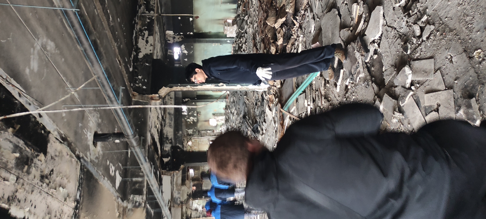 The image depicts an interior scene of a heavily damaged building, viewed from a slightly elevated perspective. The room shows extensive destruction, likely from a fire, with charred and crumbling walls, ceilings, and a floor covered in various debris such as broken concrete, tiles, and rubble.
In the middle ground, a man stands upright, facing towards the right side of the frame (if the image were rotated to standard orientation). He wears a dark jacket or suit, dark trousers, and white gloves, with dark hair and glasses. He appears to be looking down, possibly inspecting the damage on the floor or a specific detail in the immediate vicinity.
In the foreground, partially obscuring the lower left side of the image, is the back of another person's head and shoulder, wearing a dark jacket. This person is likely the photographer or an accompanying individual. Faintly visible in the background, behind and to the left of the central man, are blurry figures, some appearing to wear blue outerwear.
The ceiling features exposed pipes and wires, including blue and grey conduit lines, indicating an industrial or commercial setting rather than a residential one. A green hose or cable lies across the debris-strewn floor near the central figure. The overall impression is one of a post-disaster site undergoing examination or assessment. There is no visible text. The lighting suggests an indoor setting, possibly with some artificial light sources.