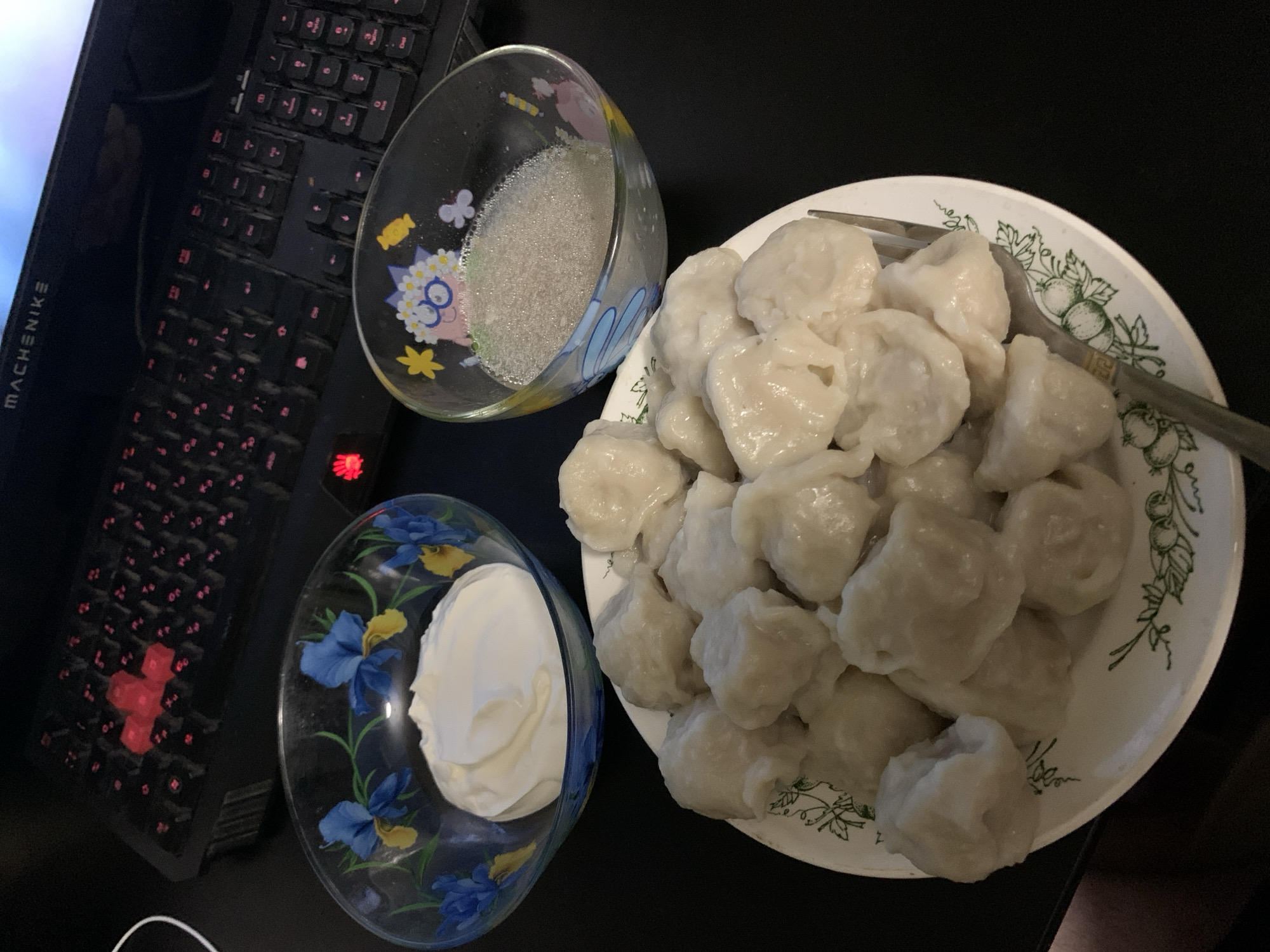 A still life image shows a plate of dumplings, a bowl of sour cream, and a bowl of sugar. A keyboard is visible in the background. The scene appears to be set indoors, on a dark surface. There is no indication of the location being Desyatikhatka, Russia.