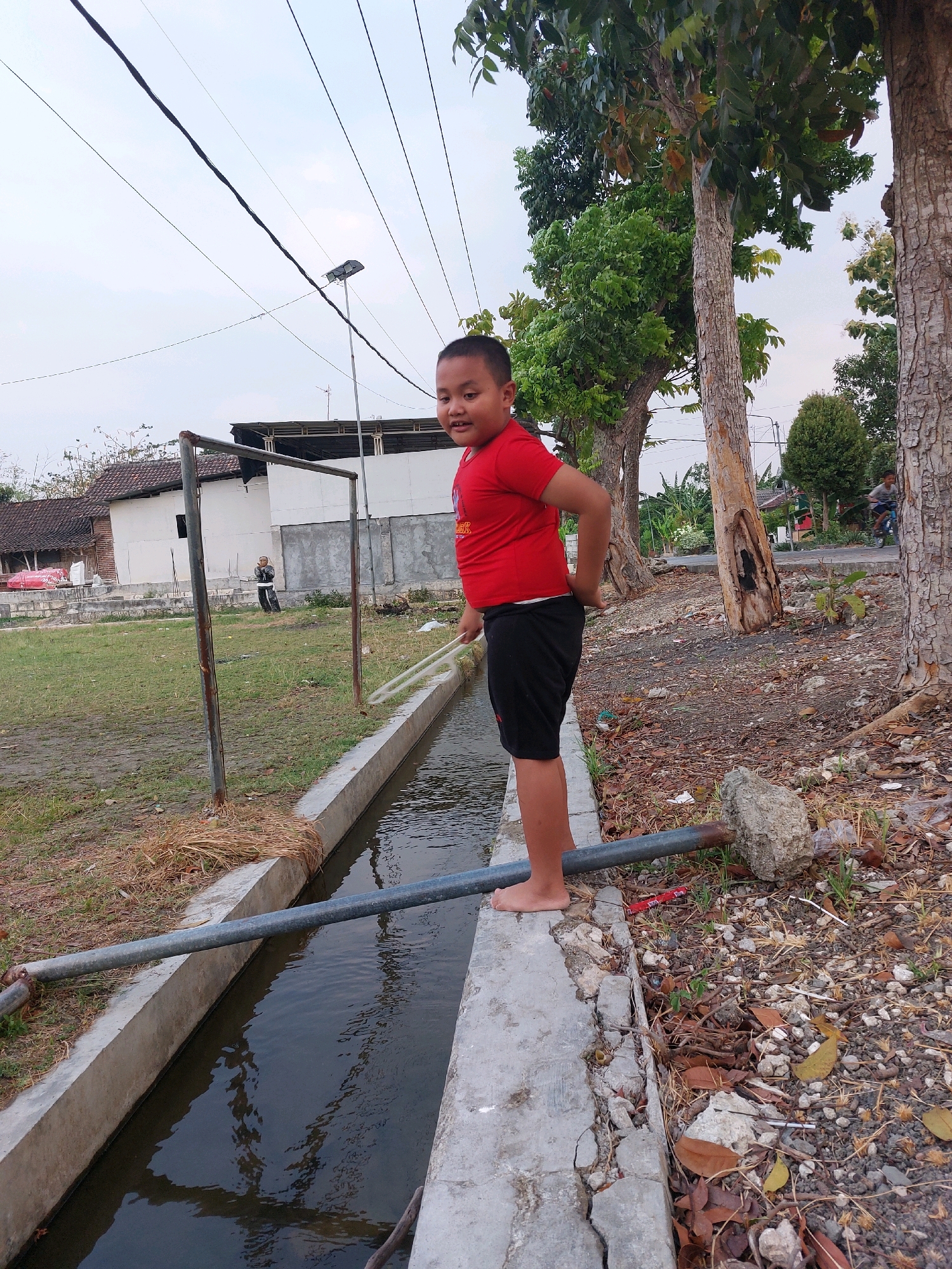In SARANGAN, Indonesia, a young boy wearing a red t-shirt and black shorts stands barefoot on the concrete edge of a water channel. He is positioned in the right half of the frame, facing towards the camera with his left hand on his hip and his right hand holding a white pole. The water channel runs along the bottom of the image, appearing dark and reflective. The area surrounding the channel is covered with dry leaves, gravel, and some sparse green vegetation. A metal pipe runs horizontally across the water channel, supported by a concrete block on the right side. In the background, to the left, a grassy field is visible with some structures, including a white building with a dark roof and a metal frame resembling a goalpost. Further in the distance, trees and a street with a person on a bicycle are present. Overhead, power lines stretch across the sky, which is overcast and light.