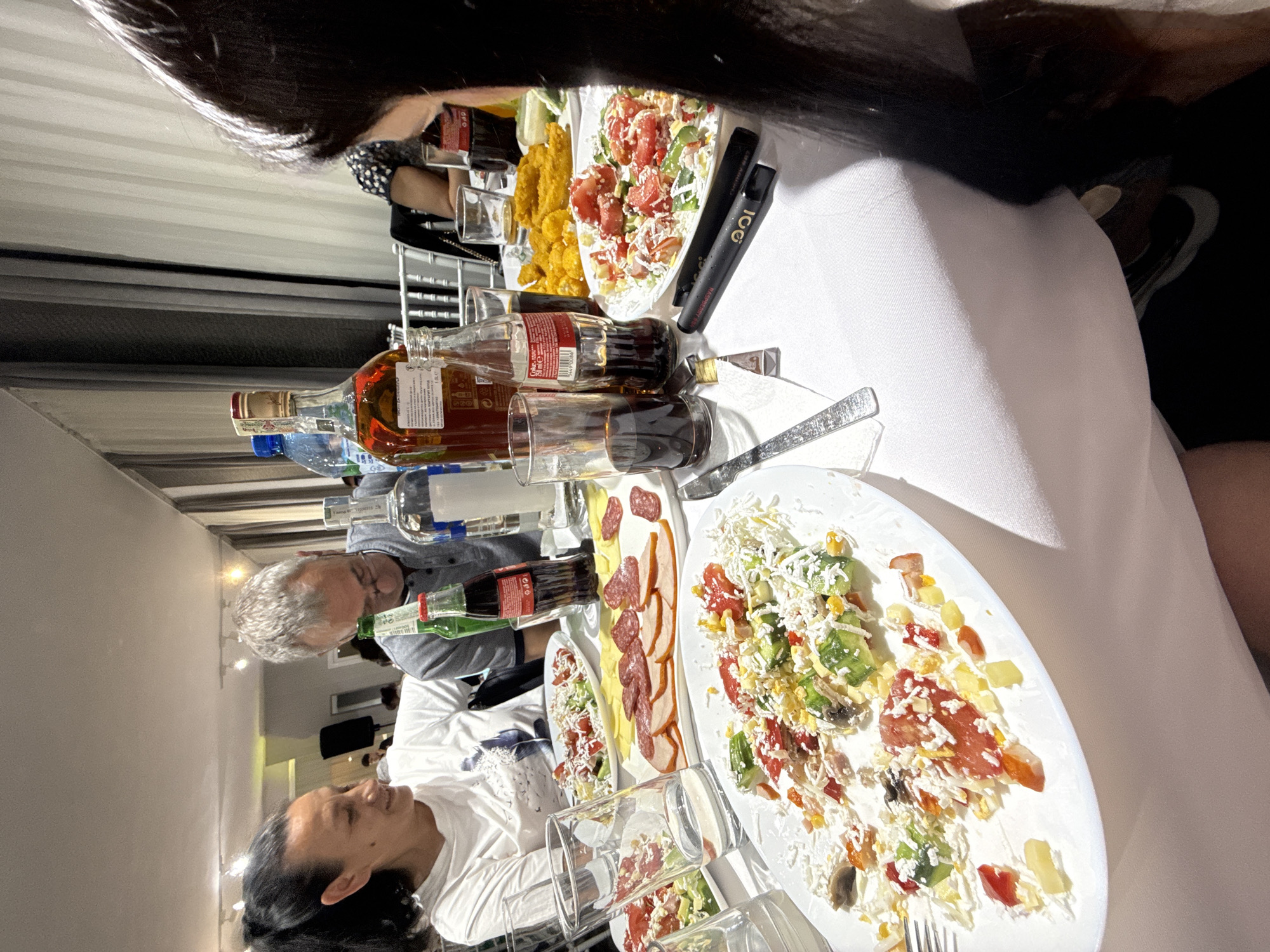 A group of people are seated around a white tablecloth-covered table laden with food and drinks, likely at an indoor gathering or event in Rogosh, Bulgaria. The food consists primarily of various salads, some appearing to be a mix of tomatoes, cucumbers, and cheese, and platters of cold cuts like ham and salami. Beverages include bottles of Coca-Cola, a clear spirit, and possibly water. The individuals present are captured in a moment of dining and conversation. One woman with dark hair pulled back in a ponytail is seated on the left, smiling and looking towards the center of the table. A man with graying hair and glasses is seated across from her, looking down at the table. Another person, whose face is not fully visible, is seated on the right. The background shows what appears to be a dining hall or event space with lighting fixtures and curtains. The overall scene depicts a social gathering focused on food and interaction.