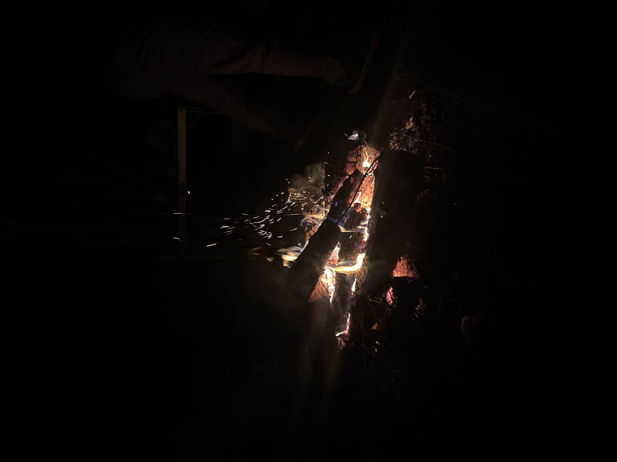 The image shows a dark, possibly nighttime scene of a burning campfire. The fire consists of several logs, with flames and sparks emanating from the wood. The area surrounding the fire is largely dark, but the fire provides enough illumination to see a person or persons sitting near the fire with what appears to be a wooden structure behind them. The photograph was taken outdoors at night in Папярнянскі сельскі Савет, Belarus.