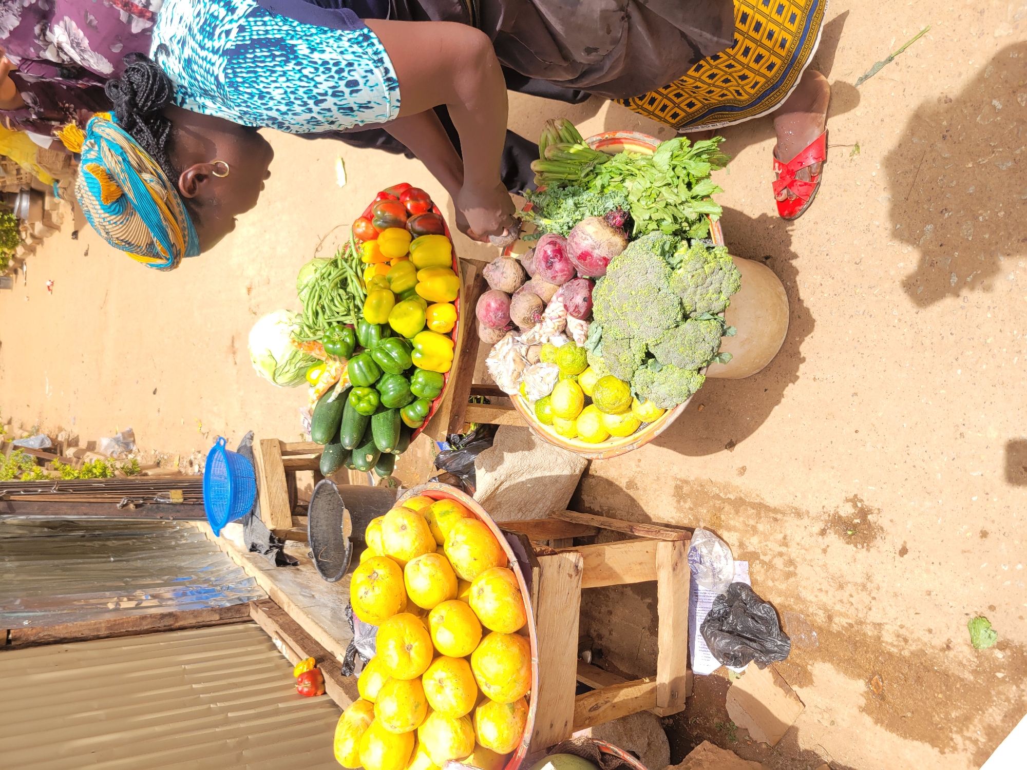 In Jos, Nigeria, an overhead shot captures a marketplace scene. The view is from above the vendors. Two women are visible. One woman, wearing a blue and white patterned top and a multi-colored headwrap, leans over a wooden table displaying produce. Her left ear has a gold hoop earring. The other woman is partially visible, wearing a black top and a yellow patterned skirt, with red sandals. Three baskets display an assortment of vegetables and fruits. One contains green peppers, yellow peppers, red tomatoes, green beans, cabbage, and zucchini. Another basket has beets, broccoli, parsley, lemons, and another leafy green. The third basket holds yellow fruits, likely mangoes. The produce sits on wooden tables and boxes. A blue plastic basket and a black metal bucket are seen near the tables. The ground is dirt, with some shadows cast by the vendors.