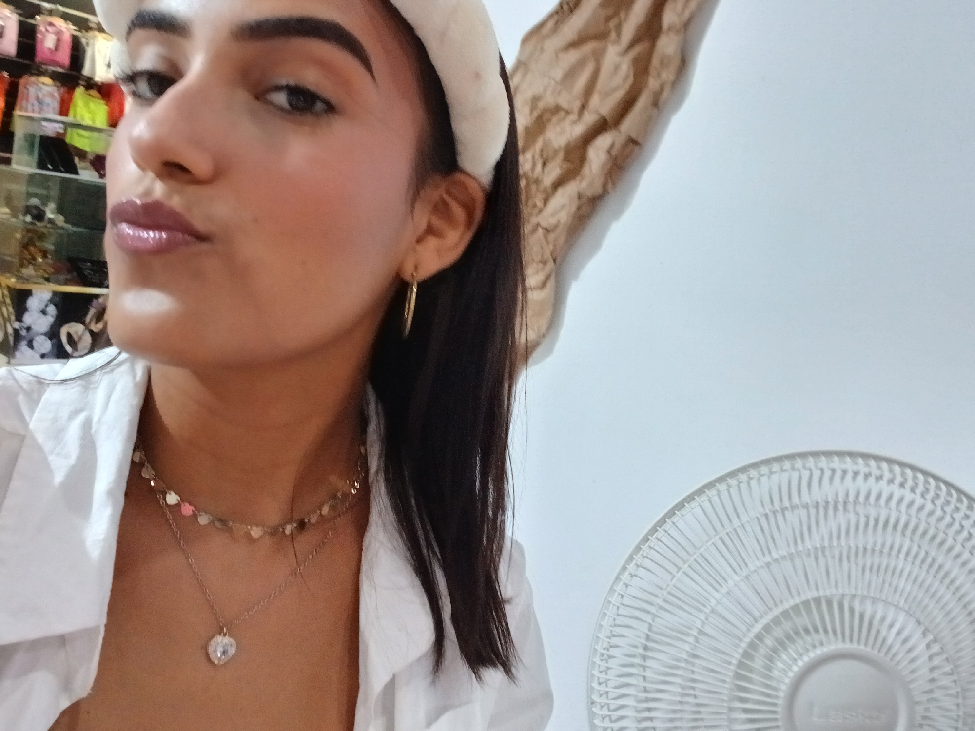 A medium shot captures a woman indoors, likely in Porlamar, Venezuela. She wears a white, collared shirt, with layered gold necklaces: one of heart-shaped charms and the other with a larger, heart-shaped pendant. A white, fuzzy headband adorns her dark hair. She has gold hoop earrings and appears to be puckering her lips. To the right, a brown crumpled paper is tacked up above a white wall and a white Lasko fan is visible in the lower right corner. In the distant background, there are clothing racks.