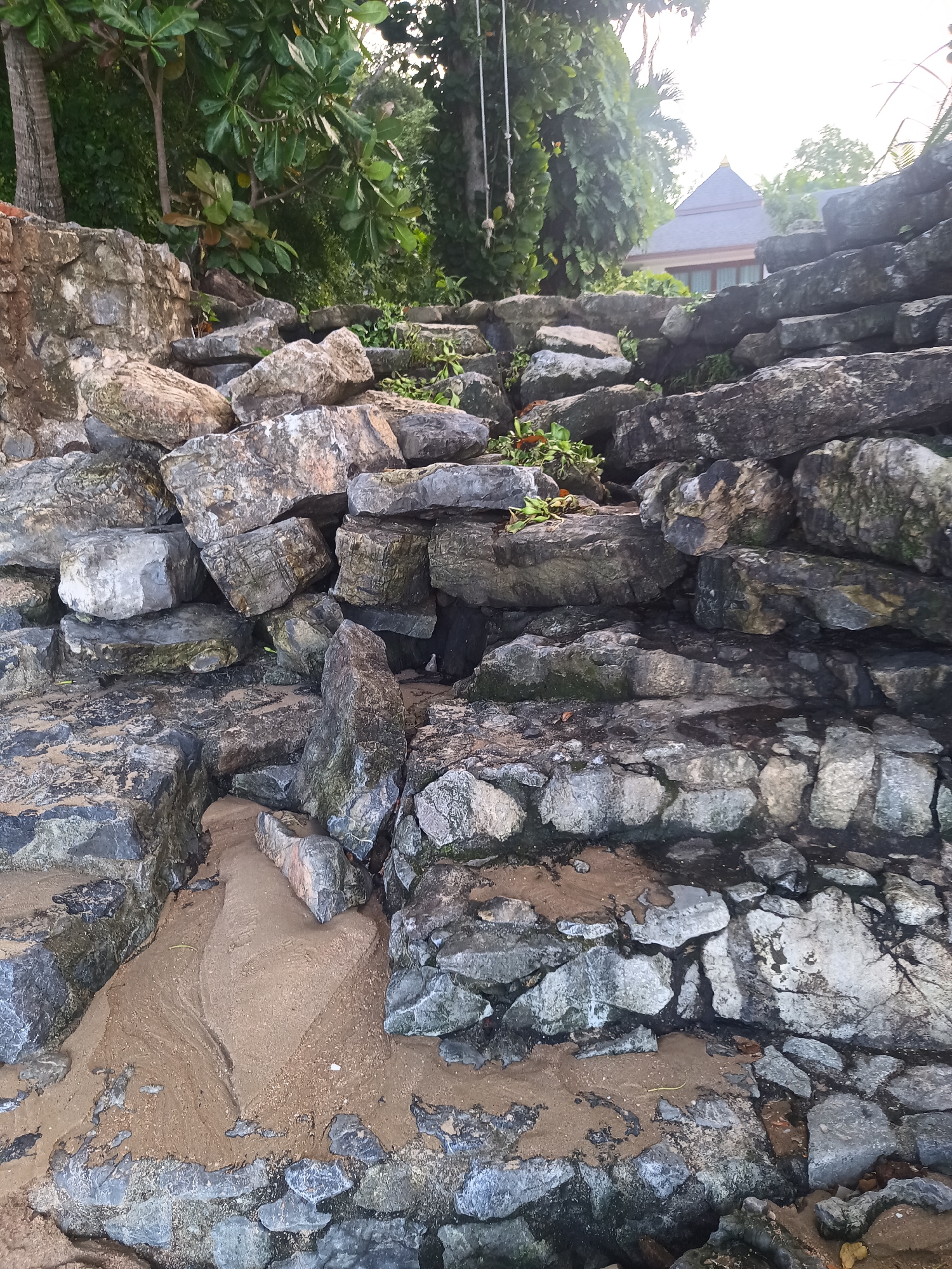 The image shows a rocky coastline in Ban Klong Haeng, Thailand. A wall is constructed from large, grey rocks, partially covered in sand. Vegetation grows on and behind the wall, with some trees visible. A structure with a pointed roof can be seen in the background. Two strands of rope are suspended from the trees.