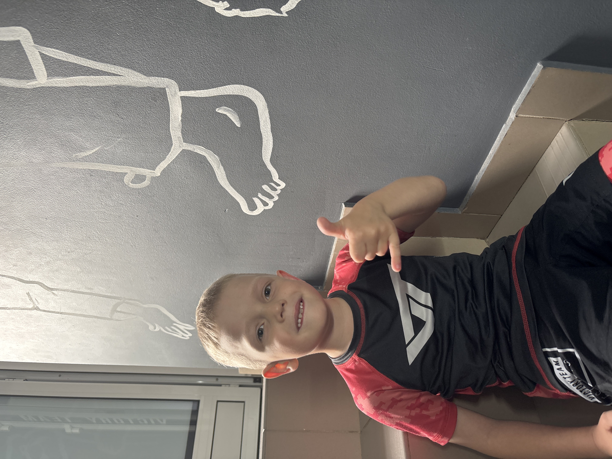 A young boy with short blonde hair and fair skin is depicted smiling broadly at the camera, making a thumbs-up gesture with his right hand while simultaneously pointing downwards with his index finger. He is wearing a long-sleeved athletic shirt (rashguard style) that is black with red sleeves and a red camouflage pattern. A large white stylized 'A' or 'V' symbol is visible on the front of his shirt. He is also wearing black athletic shorts, with "VICTORY TEAM" printed vertically in white on the side.
The setting appears to be indoors. Above and behind the boy is a dark gray ceiling or wall adorned with white line drawings of human body parts, including a detailed foot with toes, a leg, and an arm extending into a hand with fingers. The drawings are stylized and cartoon-like. To the left of the boy, part of a white-framed window is visible, suggesting natural light is present. To the right and beneath him, light brown or beige surfaces, possibly furniture or architectural elements, complete the background. The overall impression is cheerful and possibly taken in a recreational or sports-related environment. The boy's expression is happy and engaging. The location context provided is Votkinsk, Russia.