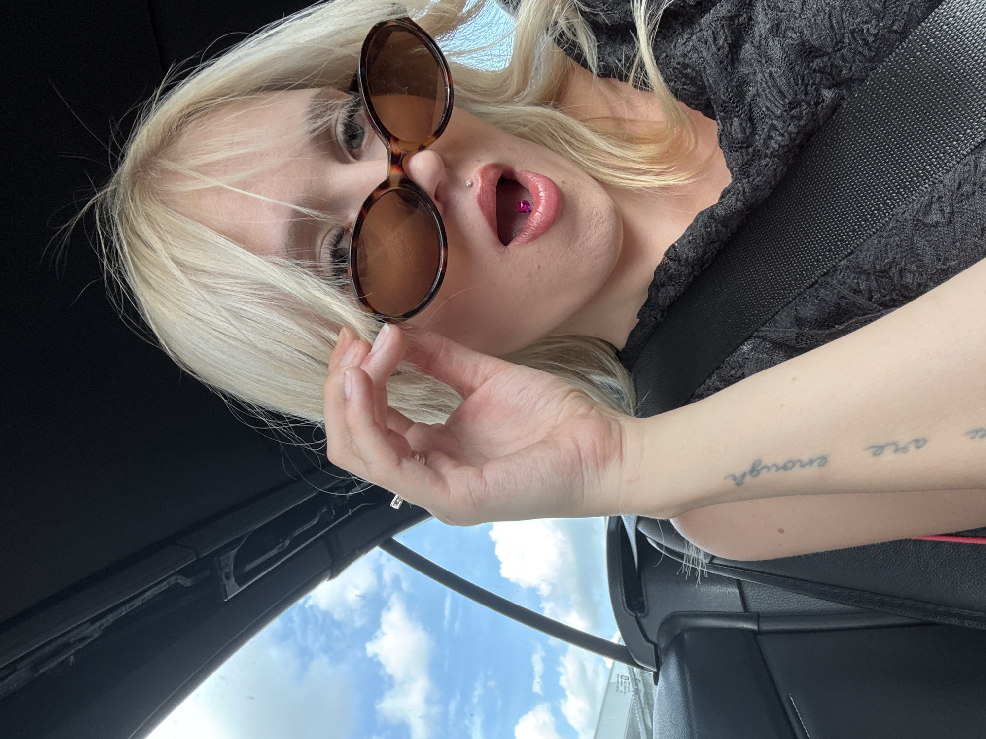 A young white woman with blonde hair and sunglasses is in a car. She is wearing a black lace top and a seatbelt. Her mouth is open, revealing a pink jewel in her mouth. She has a tattoo on her arm that says "you are enough." The sky with clouds is visible through the car window.