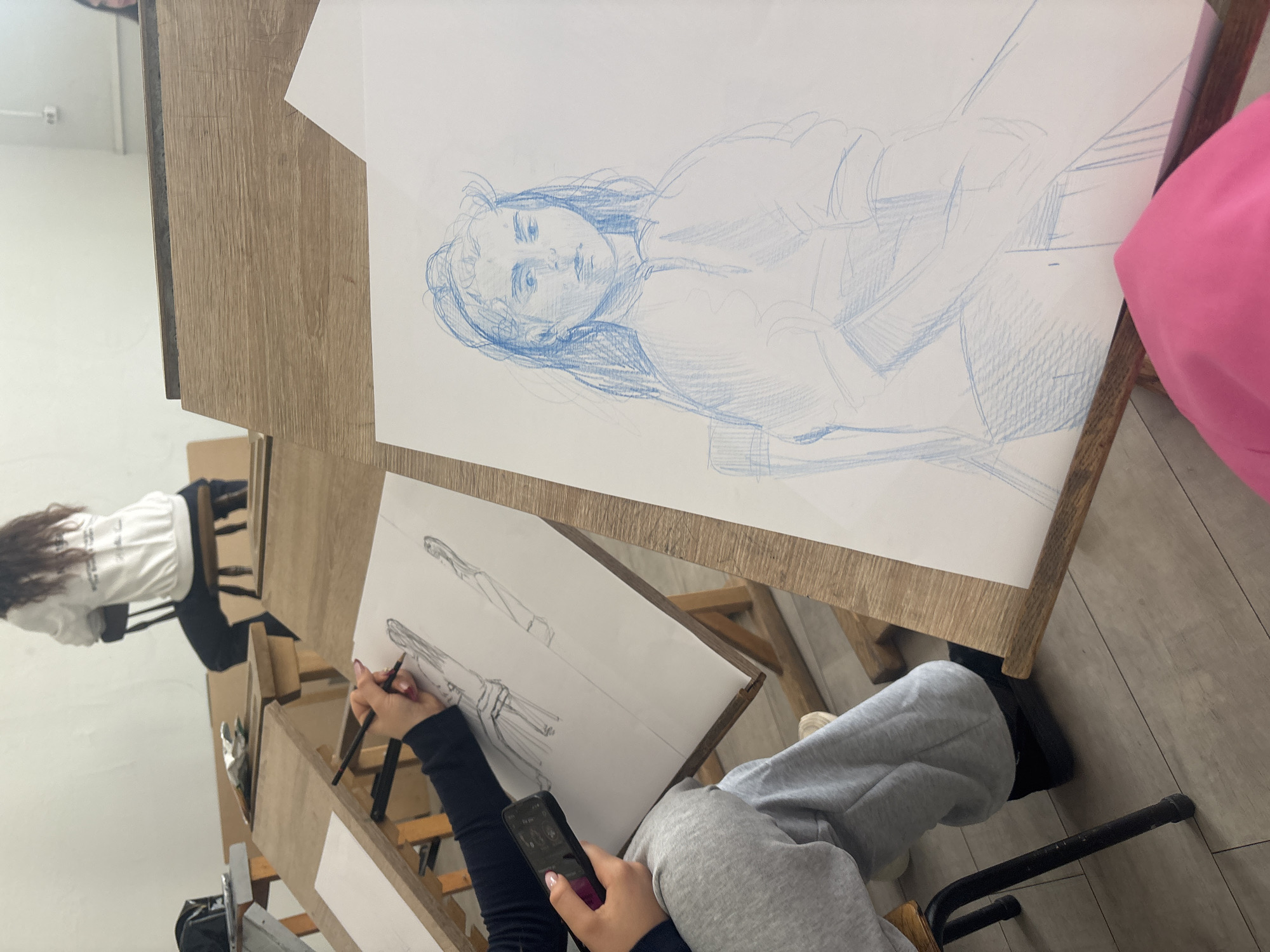 The image captures an art studio or classroom scene in Plovdiv, Bulgaria, featuring two individuals engaged in artistic activities. The setting includes multiple wooden easels or drawing desks, light-colored walls, and a floor with wood-effect tiles.
In the foreground, a person is seated, partially visible from the chest down, wearing grey sweatpants and a black long-sleeved top. Their right hand holds a pencil, actively sketching on a sheet of white paper placed on a wooden easel. This drawing is a detailed pencil sketch of a standing figure, characteristic of a fashion illustration. The person's left hand holds a black smartphone with a screen displaying various icons.
Above and behind this primary artist, a larger sheet of white paper is positioned on another wooden surface. This paper displays a blue monochrome line drawing of a person's head and upper body, executed in a loose, expressive style, possibly a portrait or life drawing study. To the far right, a vibrant pink soft object, perhaps a bag or cushion, is partially visible.
In the background, on the left side of the frame, another person is seated on a chair at a wooden desk. They are viewed from behind, wearing a white long-sleeved top with the text "Creative Studio" printed in dark letters across the back. This person has long, dark hair. An electrical outlet is visible on the wall in the upper left corner of the room. The scene suggests a daytime art class or workshop environment, with participants focused on their drawing tasks.