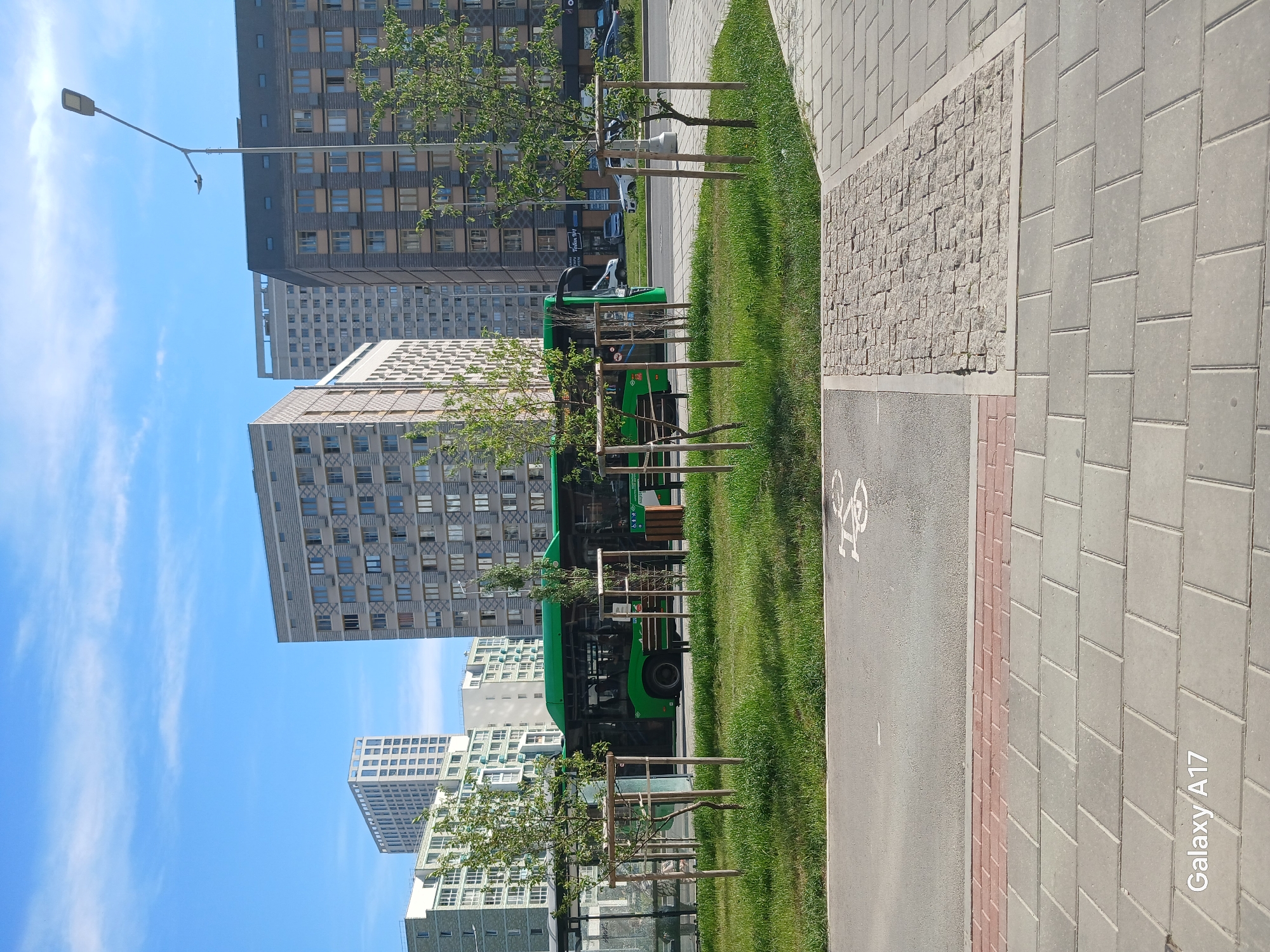 The image shows a cityscape in Yekaterinburg, Russia. In the foreground, there's a paved area with a bike lane marked by a bicycle symbol. To the right of the bike lane, there's a sidewalk constructed from rectangular gray pavers with a section of small cobblestones separating them. A strip of green grass with small trees runs parallel to the bike lane.
Behind the grass and trees, a green bus is parked along the street. In the background, there are several tall buildings with a grid-like pattern of windows, suggesting apartments or offices. A street light is visible in the upper left corner. The sky is blue with scattered white clouds. The overall scene depicts a sunny day in an urban setting, showcasing a public transportation vehicle (bus) and infrastructure for both pedestrians and cyclists.