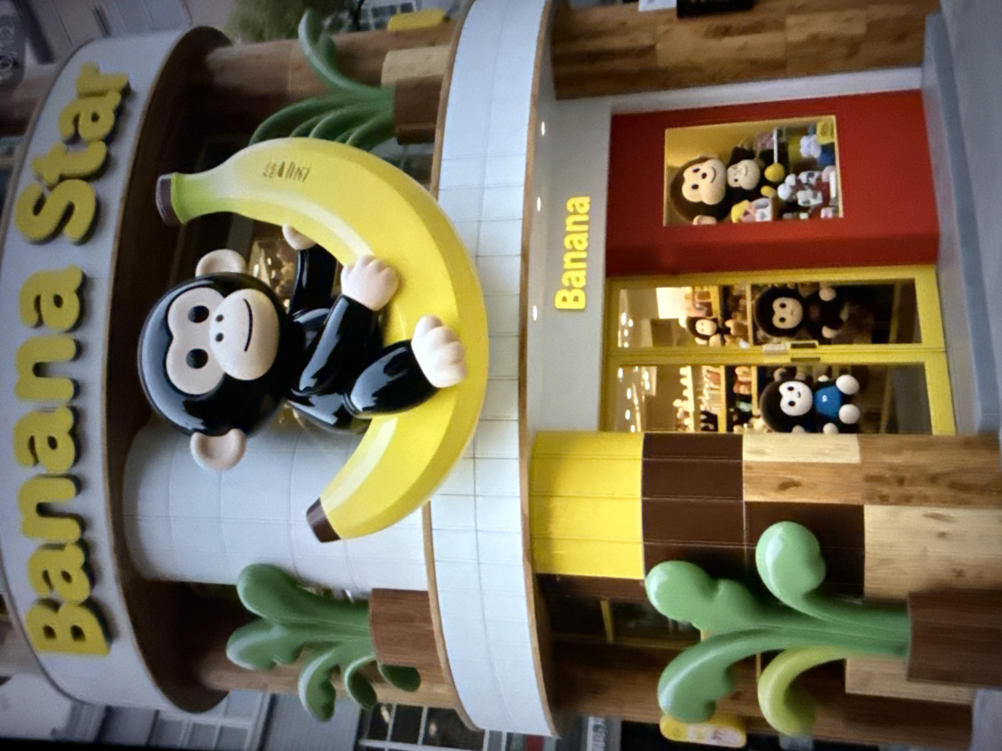 This image shows the exterior of a themed store called "Banana Star" located in Taipei, Taiwan. The store's facade is decorated with a large, cartoonish monkey character sitting on a giant banana. The store's name is displayed prominently in yellow lettering above the entrance. The building features a modern architectural style with rounded elements and a mix of bright colors, including yellow, red, and brown. The entrance is a glass storefront with yellow doors, revealing shelves stocked with plush toys, many of which are also monkey-themed. Green, stylized plant decorations are visible on either side of the entrance. The lighting suggests it is daytime or a well-lit evening. The overall impression is of a fun, playful, and inviting retail establishment.