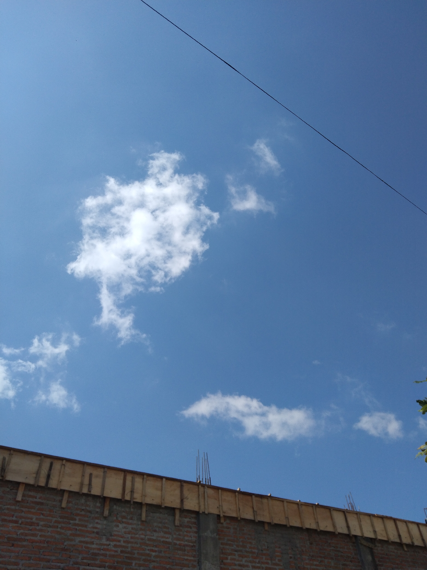 The image shows the upper portion of a brick building under construction, with the blue sky above. The building has a wooden formwork along the top edge, and rebar protruding upwards. Several white clouds are scattered across the sky, and a thin black wire runs diagonally across the upper part of the frame. The scene appears to be outdoors, possibly in a residential or urban area in Kelet, Indonesia, given the architectural style and materials. No people are visible in the image.