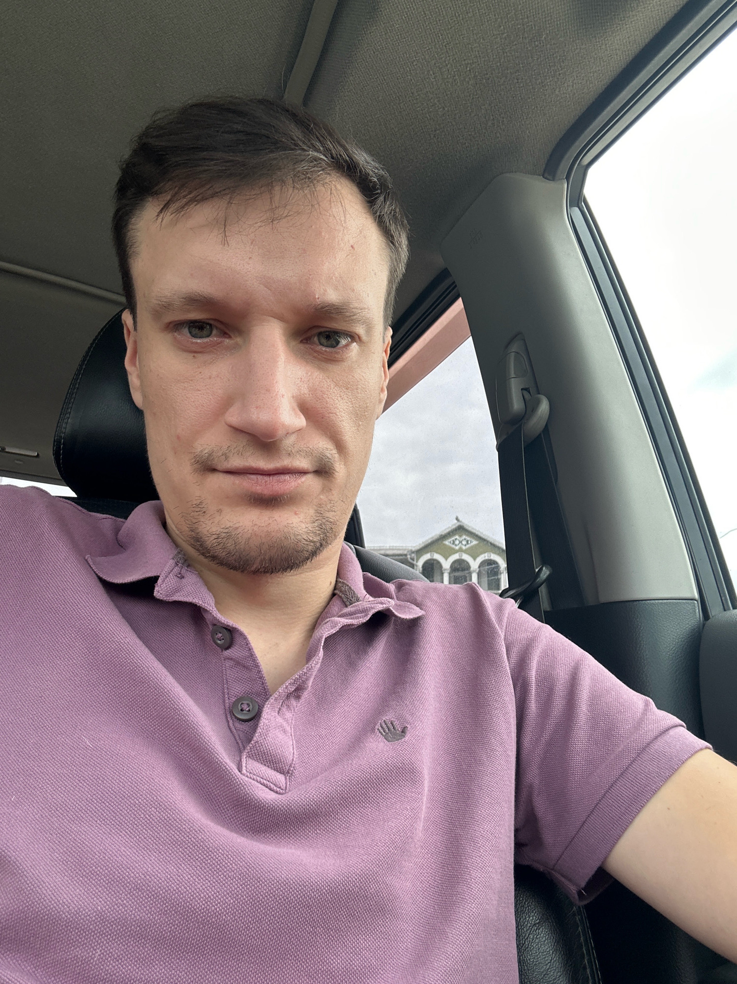A man is sitting in the driver's seat of a car, looking at the camera. He has light skin, dark brown hair, and green eyes. He is wearing a purple polo shirt. The interior of the car is visible, including the dashboard and the headliner. The window shows a cloudy sky and a building with arched windows. The car is parked on a street in Shatava, Ukraine.