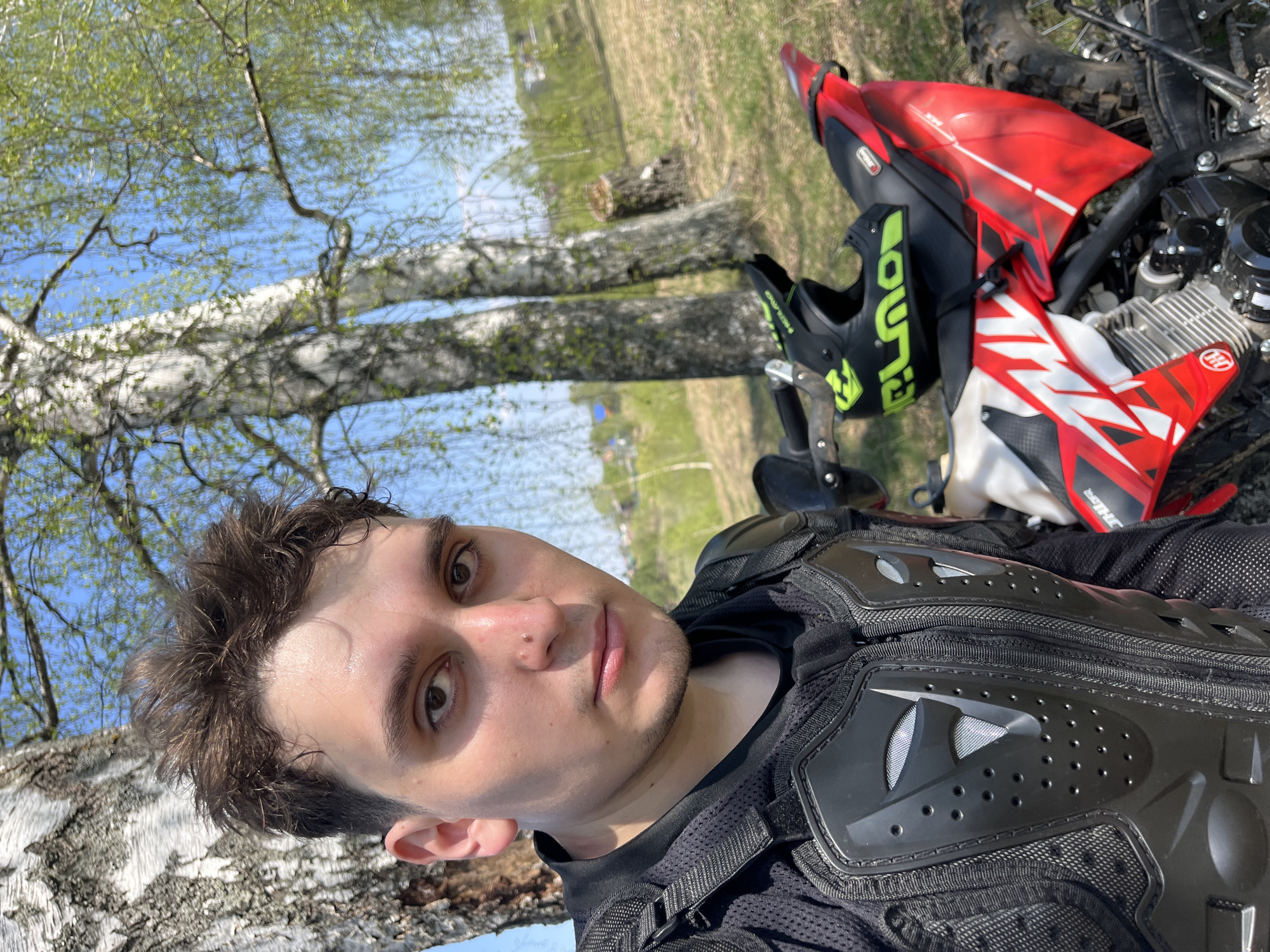 A selfie shot from a low angle captures a man wearing motocross protective gear, standing near a red and white motorcycle. The background includes birch trees with emerging leaves and an open grassy area. The image, likely taken in the town of Vasilyevo, Russia, based on the landscape characteristics.