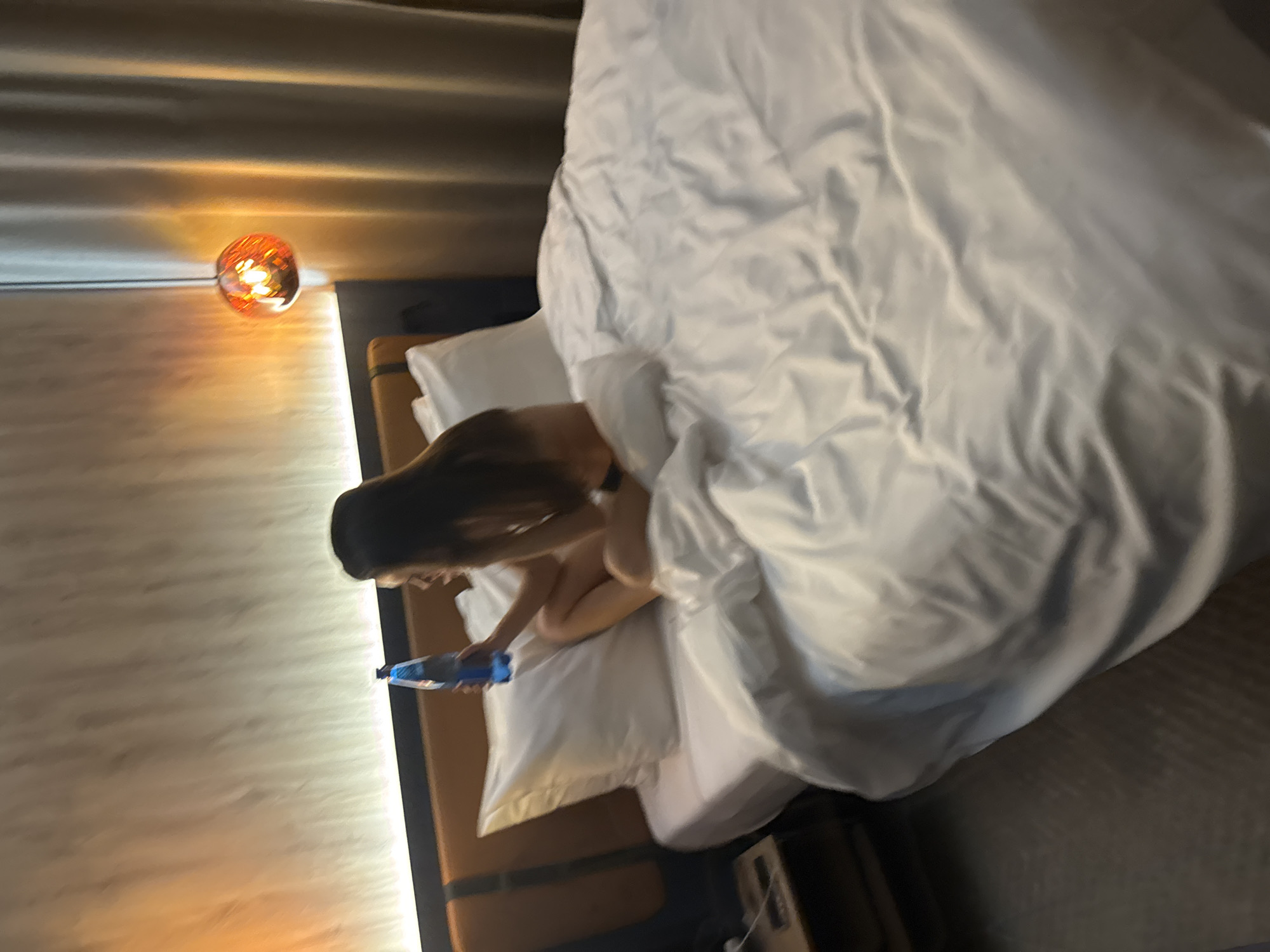 The image displays an overhead, slightly tilted perspective of a modern bedroom or hotel room, softly lit by artificial light. The primary subject is a person seated on a bed, viewed from behind and the side. They have long, dark hair and appear to be wearing a dark top, possibly a bra or bikini top, with a portion of their bare back and side visible. The person is leaning forward, looking downwards, and holding a blue bottle.
The bed is covered with a large, rumpled white duvet and equipped with white pillows. To the person's left, a bedside lamp with a distinctive spherical, amber-colored glass shade casts a warm, bright glow. The wall behind the bed features a textured panel, subtly illuminated by a linear light source that contributes to the room's cozy ambiance. A dark nightstand or the bed's base is visible in the lower right corner. The reliance on artificial lighting suggests the scene takes place during the evening or night. The person's posture conveys a sense of relaxation or quiet activity. No text is visible in the image.