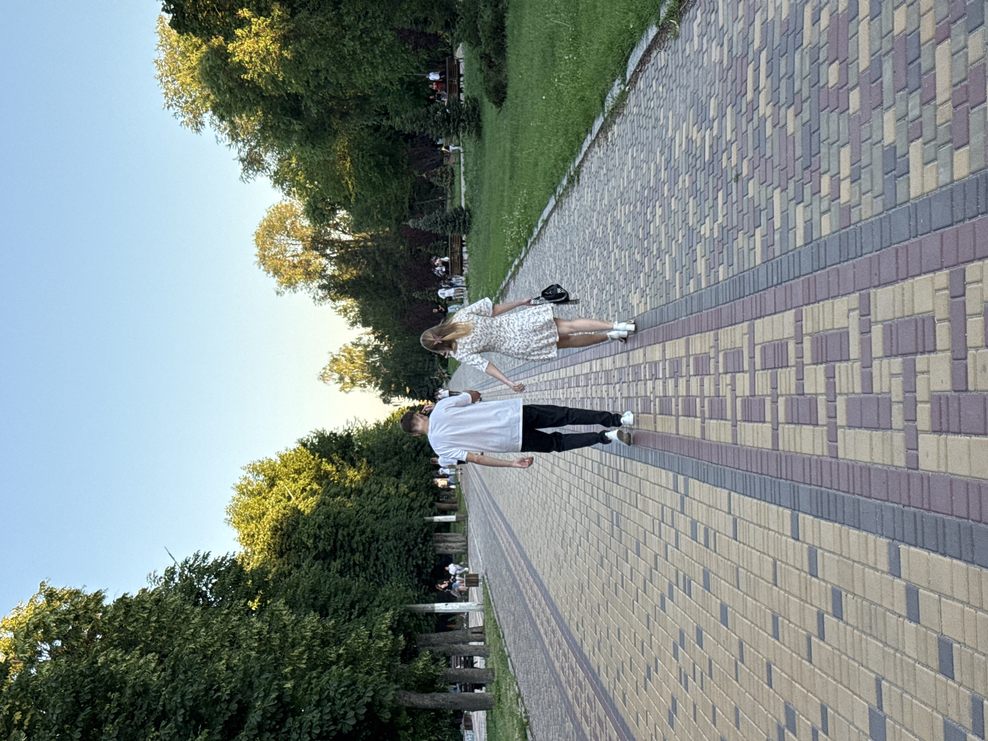 An outdoor, eye-level, full-shot photograph captures a young couple walking away from the camera, hand-in-hand, down a paved park pathway lined with trees. The pathway is constructed with tan, brown, and grey rectangular pavers arranged in a decorative pattern. To the left of the pathway, a row of mature trees with dense green foliage borders a grassy area with park benches and other people sitting and walking. To the right of the pathway, the grassy area continues, with more trees and a clear blue sky above. The man is wearing a white t-shirt, black pants, and white sneakers. The woman is wearing a short, white, floral-patterned dress and white sandals, and carries a black handbag. The couple appears to be enjoying a casual stroll in a park setting.