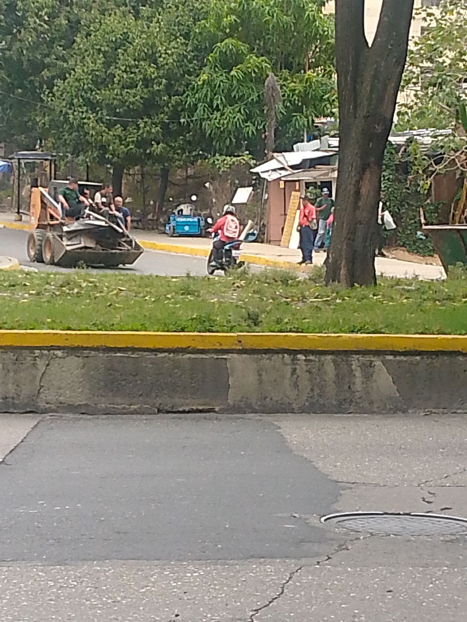The image captures a scene on a street in Caracas, Venezuela. A small, front-end loader is parked on the side of the road, occupied by at least three people. A motorcyclist wearing a red jacket and backpack is riding nearby. Several other individuals are standing on the sidewalk near a building with a temporary or makeshift structure attached to it. The environment includes trees, grass, and typical city infrastructure such as sidewalks, curbs, and a manhole cover in the road.