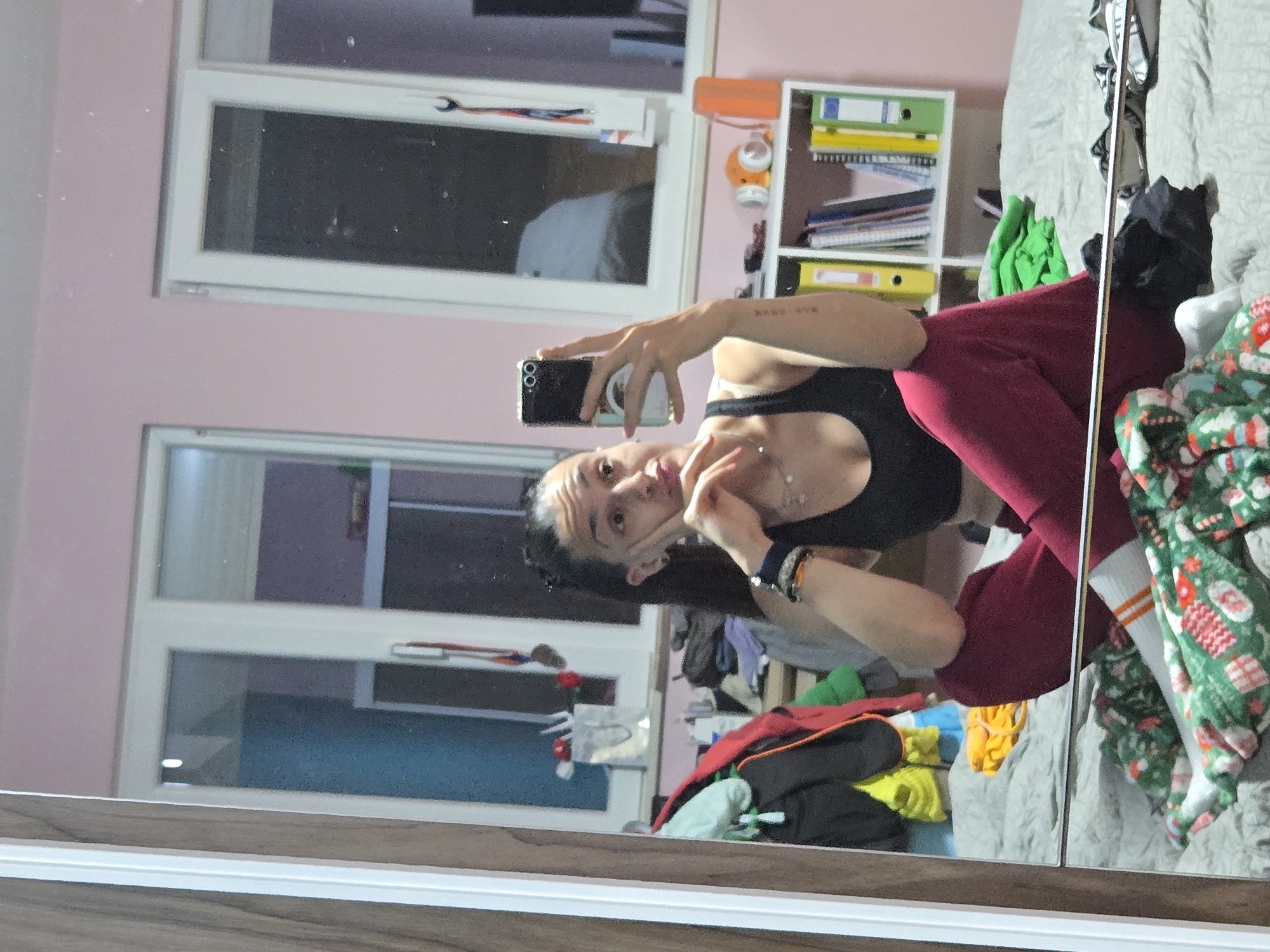 A young woman is taking a selfie in a mirror. She is wearing a black sports bra and maroon leggings. She has a dark hair pulled back. The room in the mirror appears to be a bedroom with windows and a shelf. There is a bed with multiple blankets. The location is Pleven, Bulgaria, based on the window view.