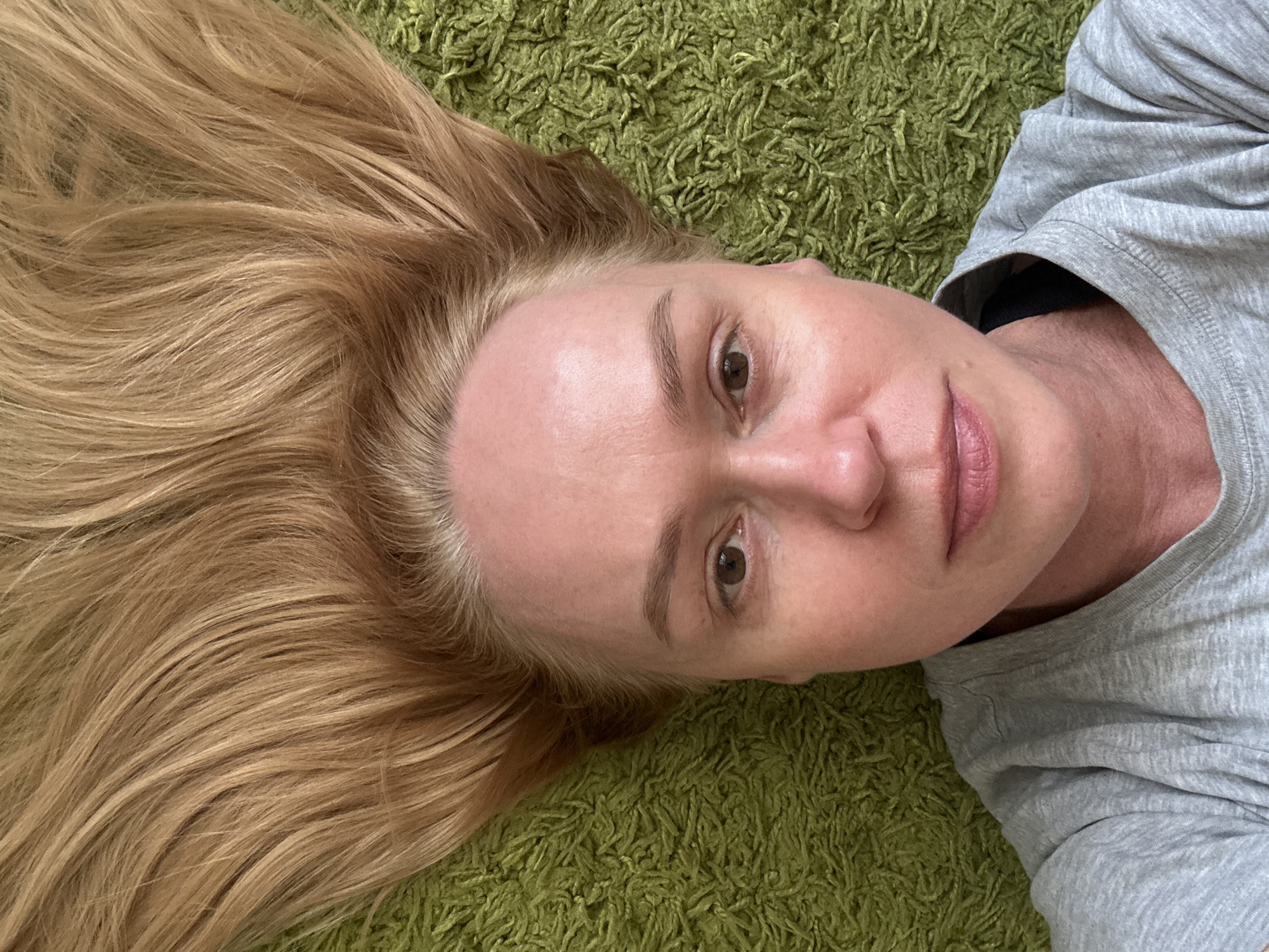 A woman with light blonde hair is lying on a green shaggy rug, possibly indoors. She is wearing a light gray t-shirt. The woman's face is visible, revealing brown eyes, fair skin, and pink lips. The background consists of the green rug, creating a textured backdrop. The image does not provide sufficient information to determine if the location is Chaikovsky, Russia. There is no specific location identified in the image.