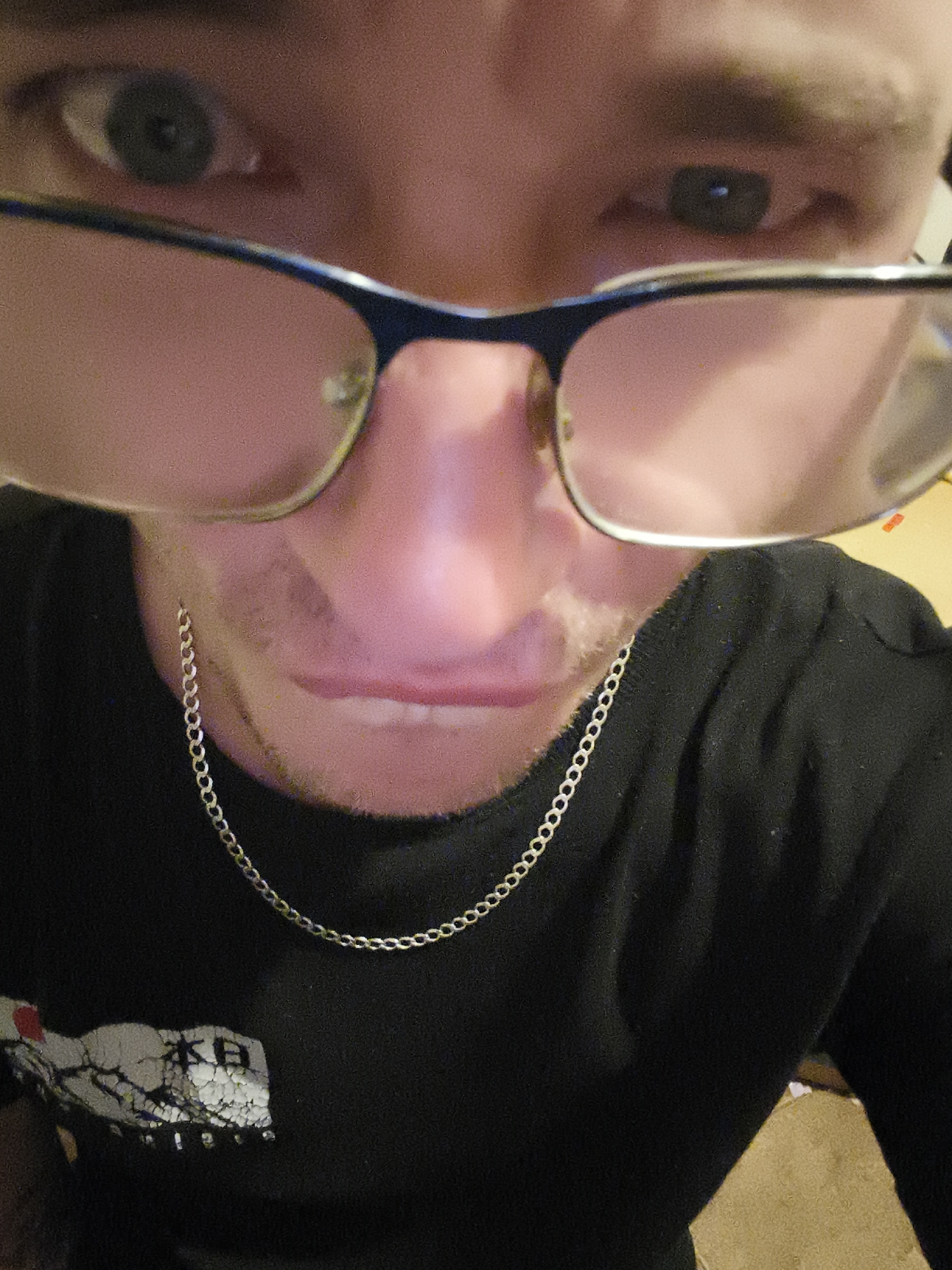 A close-up shot of a man wearing glasses and a silver chain. He is wearing a black t-shirt with a graphic on the left side. The man's face is the main focus, with his eyes, nose, and mouth clearly visible. The photograph does not contain information related to Nowy Tomyśl, Poland.