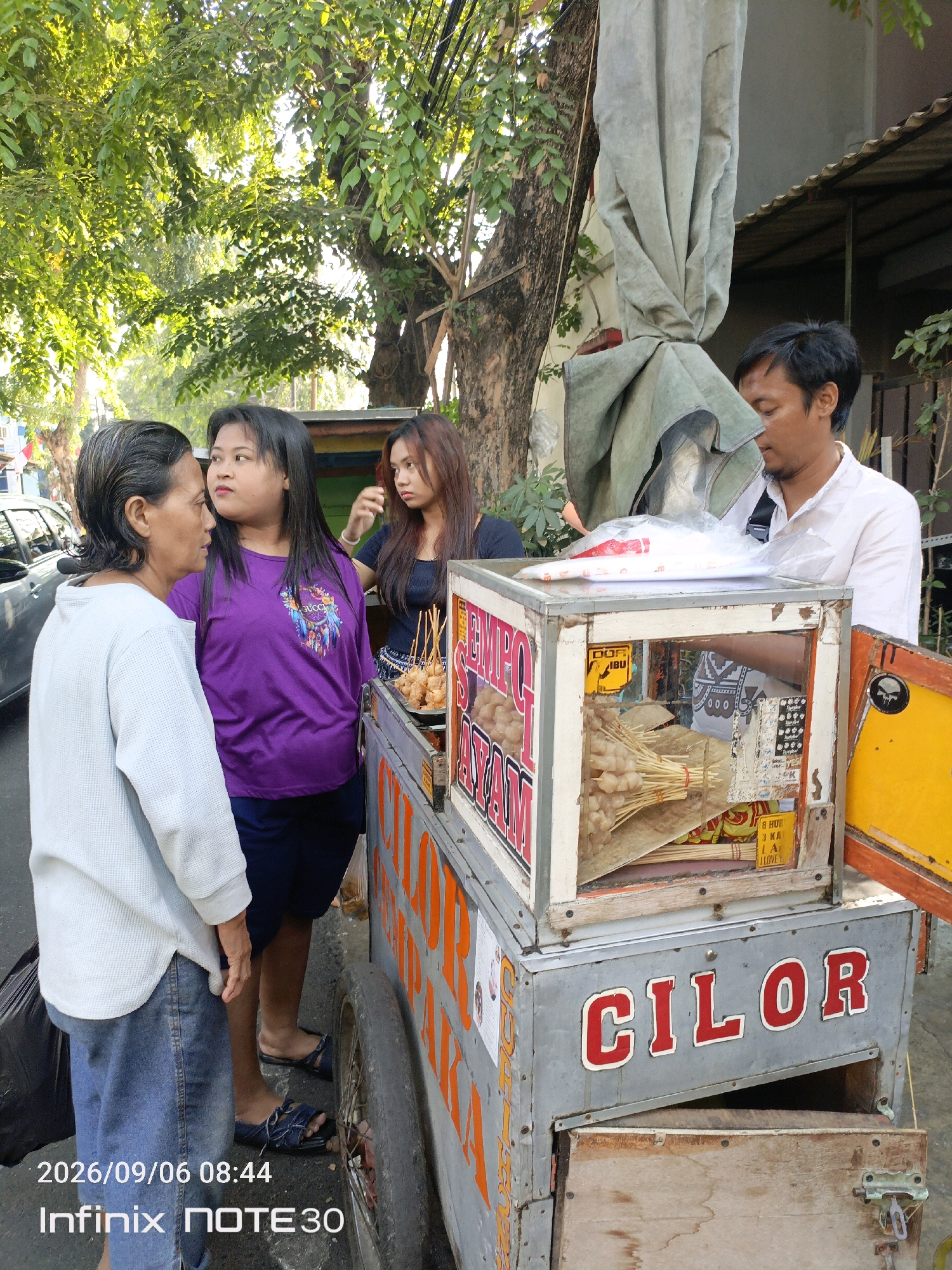 An outdoor street food stall is depicted in this image, featuring a man and three women. The man, wearing a white collared shirt, stands behind the stall, which is a wheeled cart with a glass display case filled with food items on skewers. The cart is labeled "CILOR" in large red letters. The women are standing in front of the stall, appearing to interact with each other or potentially the vendor. The woman on the far left wears a light blue long-sleeved shirt and jeans, the middle woman wears a purple t-shirt and dark skirt, and the woman on the right wears a dark top. The background shows trees, a building, and a parked car, suggesting an urban environment. The lighting indicates it is daytime, with sunlight filtering through the trees. The timestamp in the bottom left corner indicates the image was taken on September 6, 2026, at 08:44.