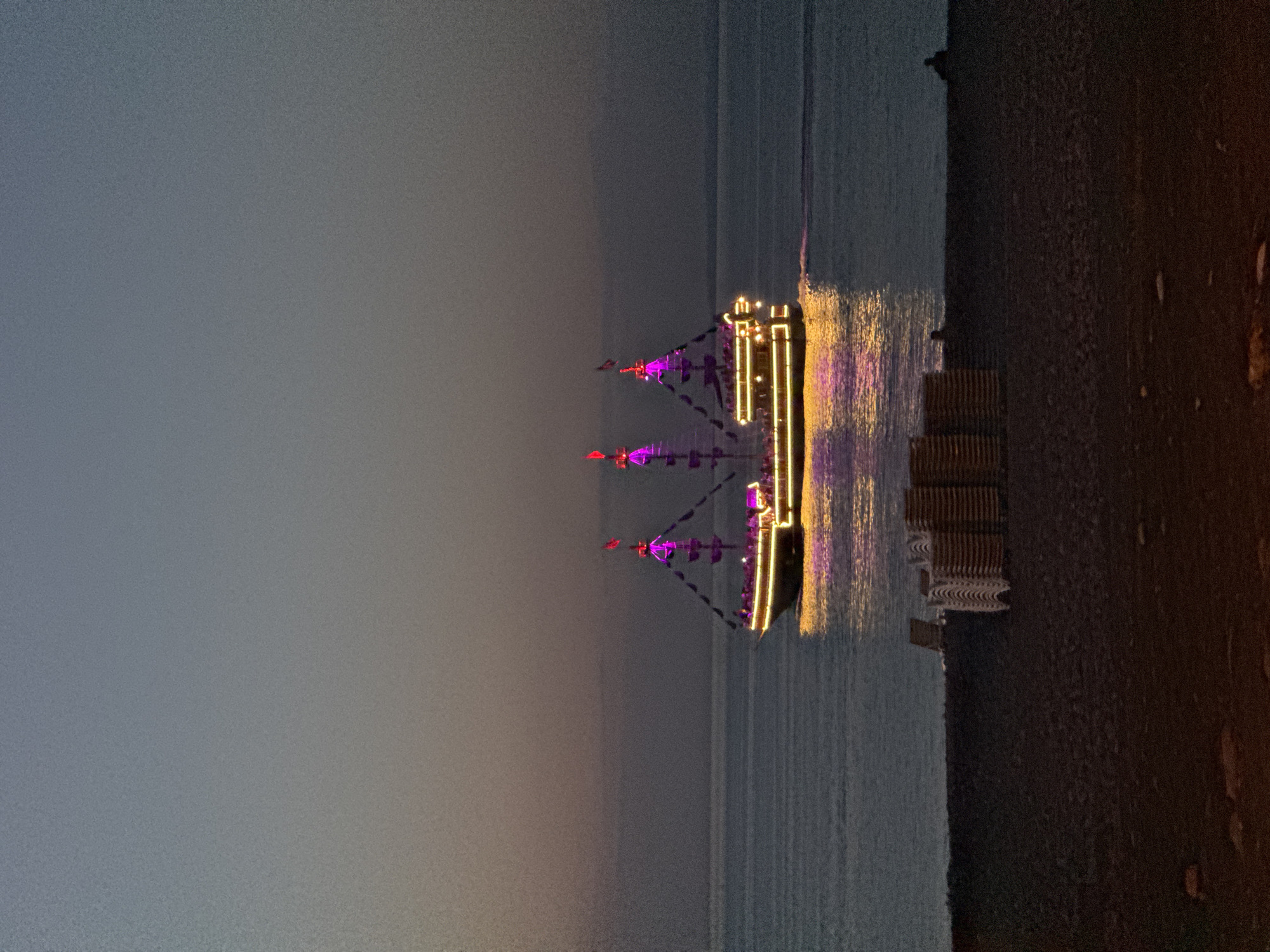 A pirate-themed boat illuminated with purple and yellow lights sails on a calm sea at dusk. The sky transitions from a pale grey near the horizon to a darker grey above. In the foreground, a beach is visible with stacked lounge chairs and two silhouetted figures sitting on the sand. The scene is likely the Batumi coastline, Georgia, given the commonality of such illuminated tourist boats operating in the Black Sea during the evening.